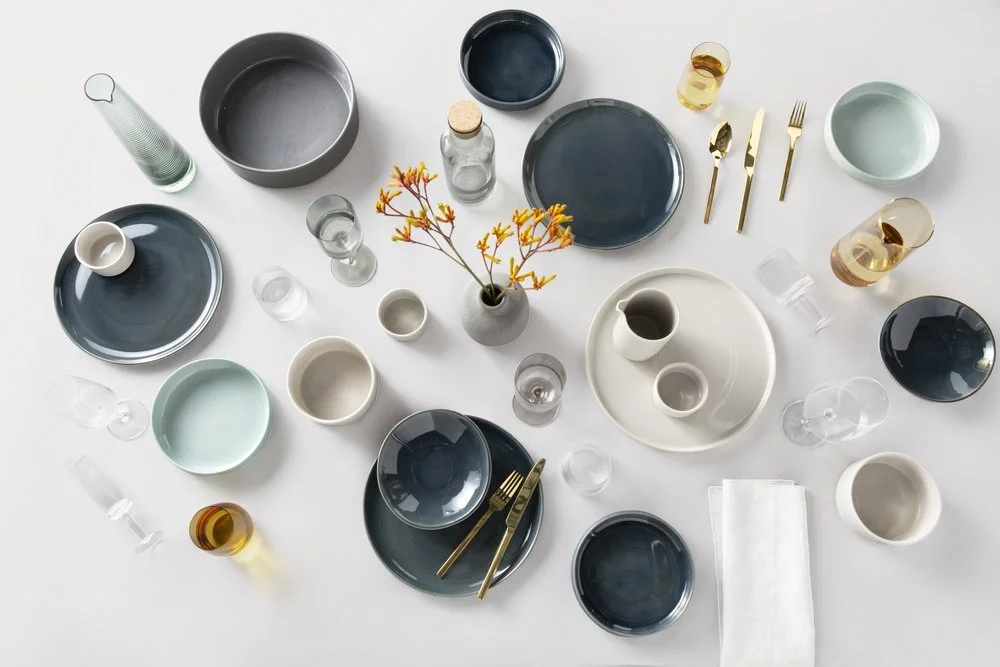 Table setting with plates, bowls, glasses, cutlery, a vase with orange flowers, and various drinks on a white surface.