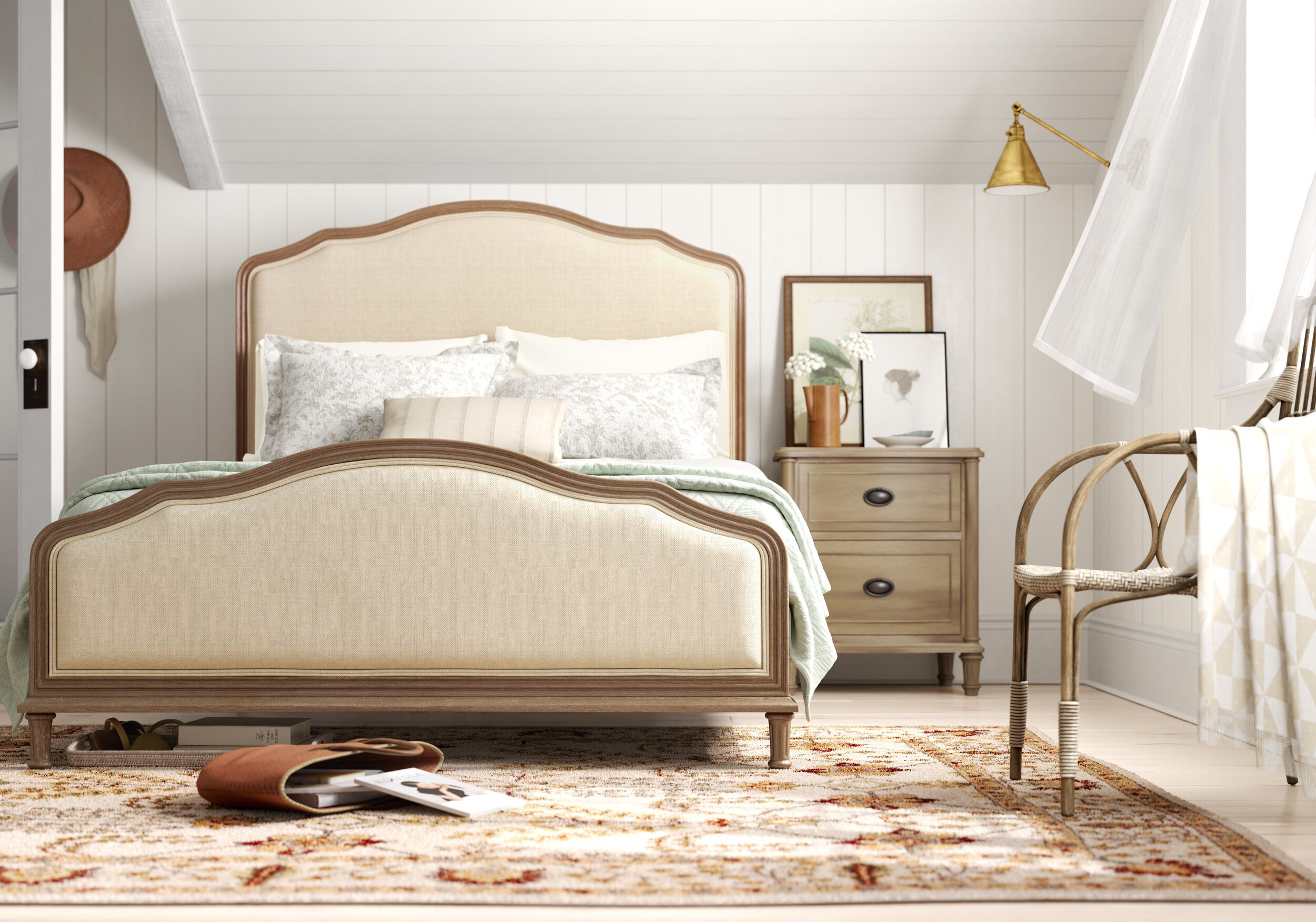 A cozy bedroom with a beige upholstered bed, a wooden nightstand with framed artwork, and a woven chair. Items on the floor include an open purse and books, with sunlight coming through a window.