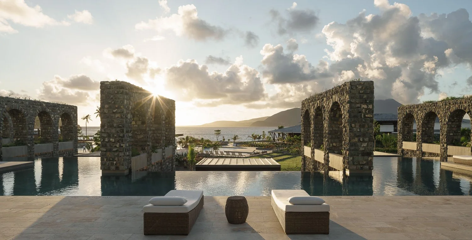 Swimming pool area with stone arches, lounge chairs, and a view of the ocean at sunset.