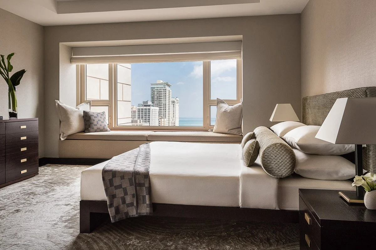 Modern hotel bedroom with a large window seat overlooking city buildings and the ocean, white bedding, pillows, and bedside lamps.