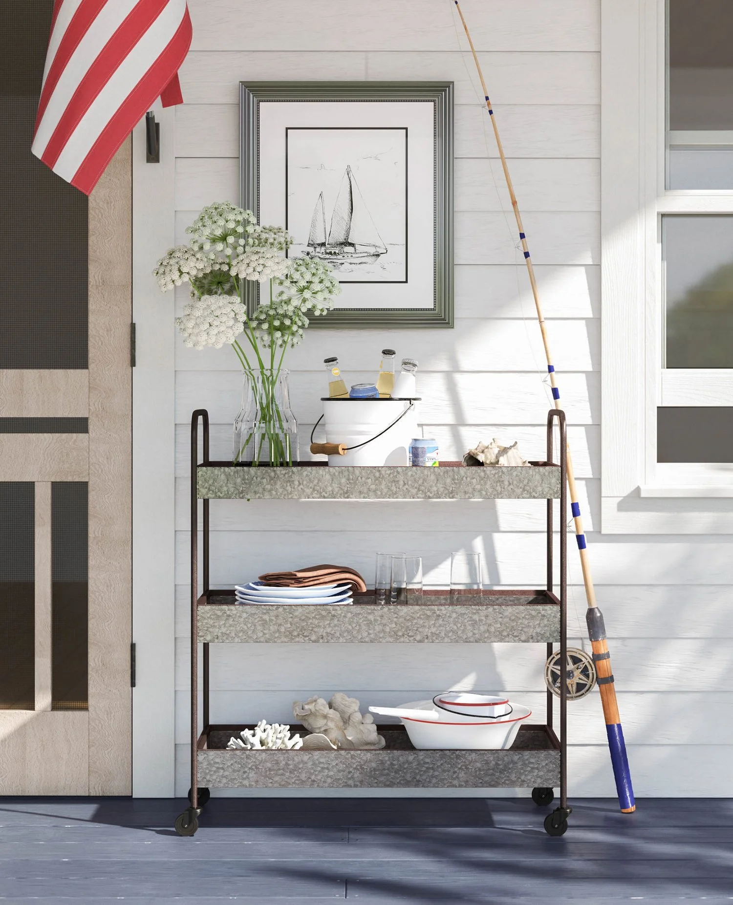 A metal cart with three shelves holds a clear vase with white flowers, a white bucket with bottles, a stack of plates and glasses, and white bowls with spoons. A fishing rod leans against the wall next to a window, and a framed black-and-white sailbo