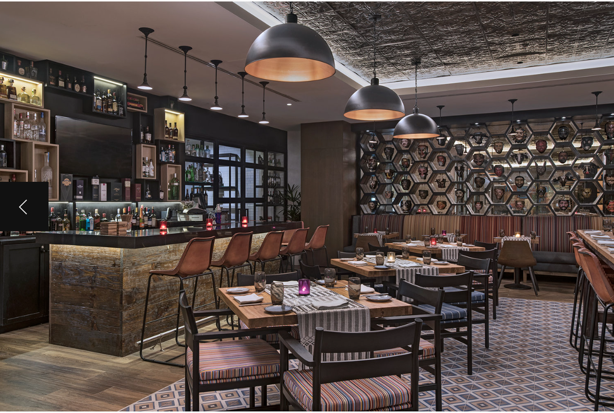 Elegant restaurant with a bar area, modern pendant lighting, and neatly set tables with candles and tableware, decorated with a honeycomb pattern wall featuring masks.