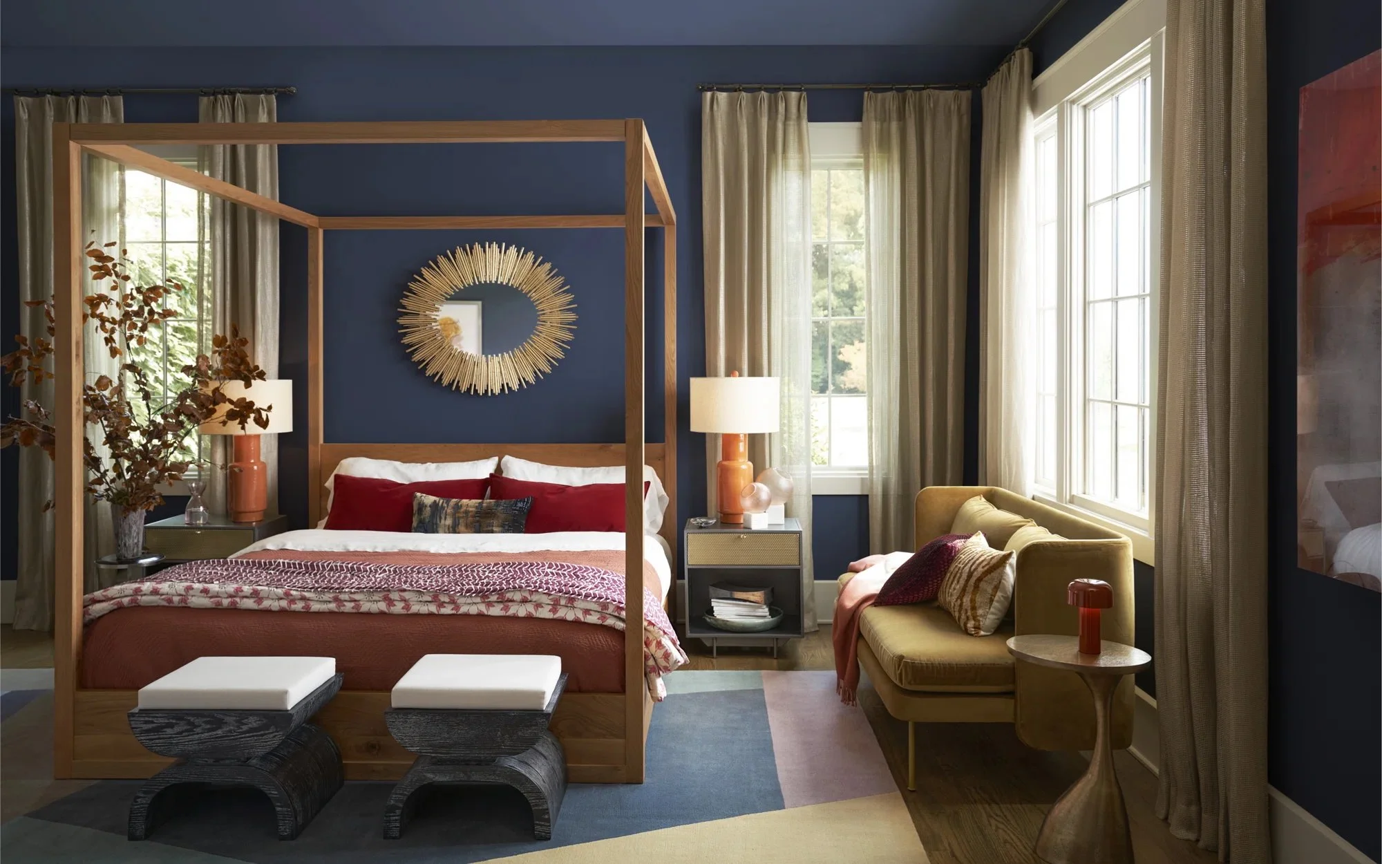 A bedroom with a dark blue accent wall behind a wooden four-poster bed with red and patterned bedding. There are two dark wooden stools with white cushions at the foot of the bed. On each side are matching nightstands with large lamps, and gold curta