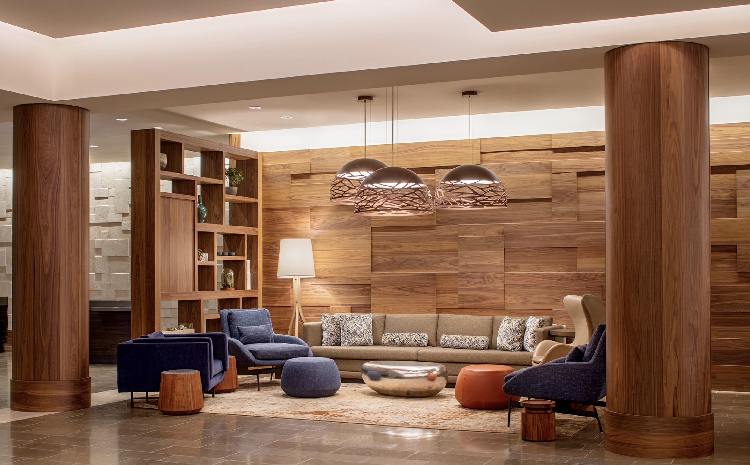 Elegant hotel lobby with wooden walls, large circular wood pendant lights, beige sectional sofa with patterned cushions, blue and beige armchairs, various side tables, and a tall floor lamp. Wood-paneled walls and columns create a warm atmosphere.