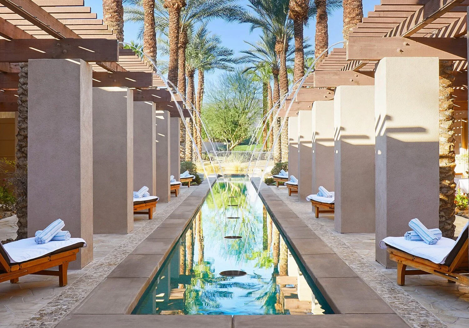 Luxury outdoor lounge area with a narrow pool, fountains, wooden lounge chairs with rolled towels, surrounded by tall palm trees and modern structures, in a sunny setting.