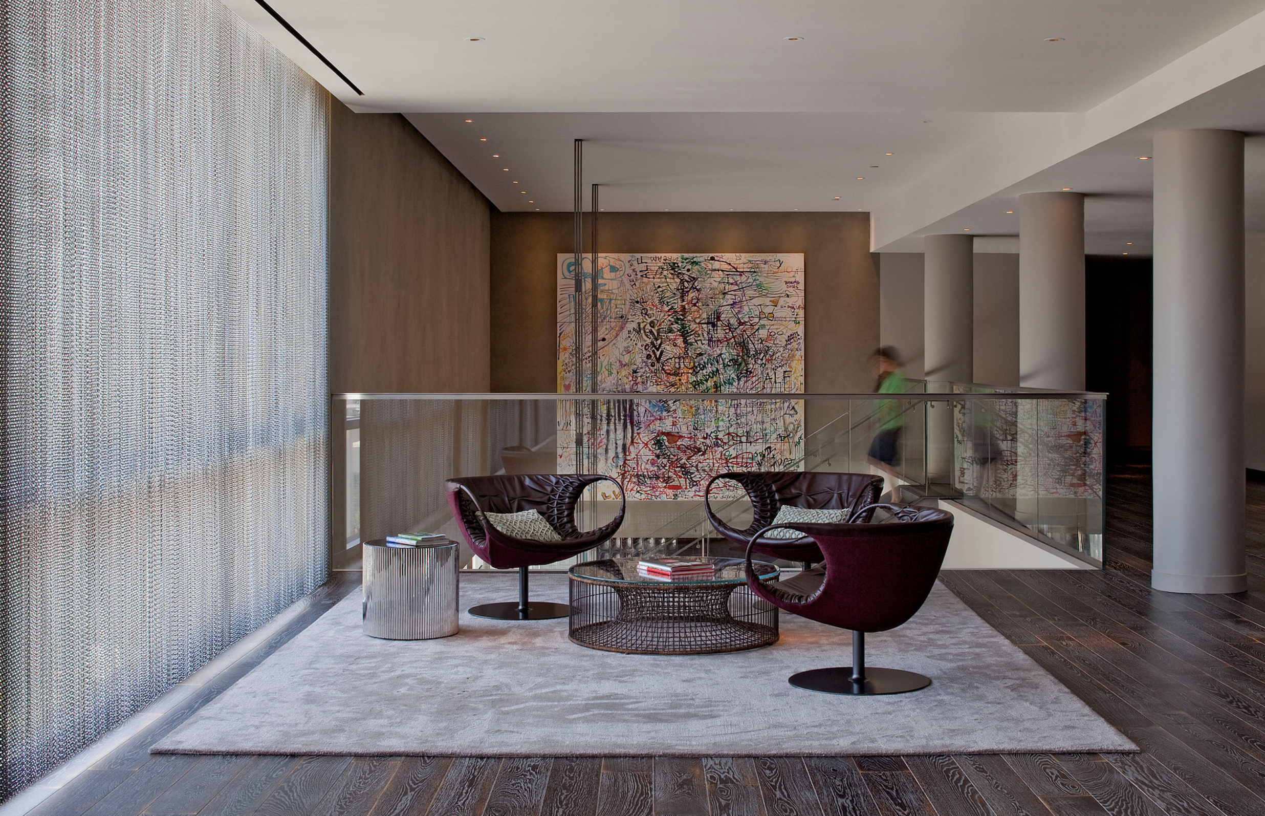 Modern lounge area with three burgundy chairs around a glass coffee table on a light-colored rug, with an abstract colorful painting on the beige wall behind, large textured window to the left, dark wooden floor, and a person walking in the backgroun