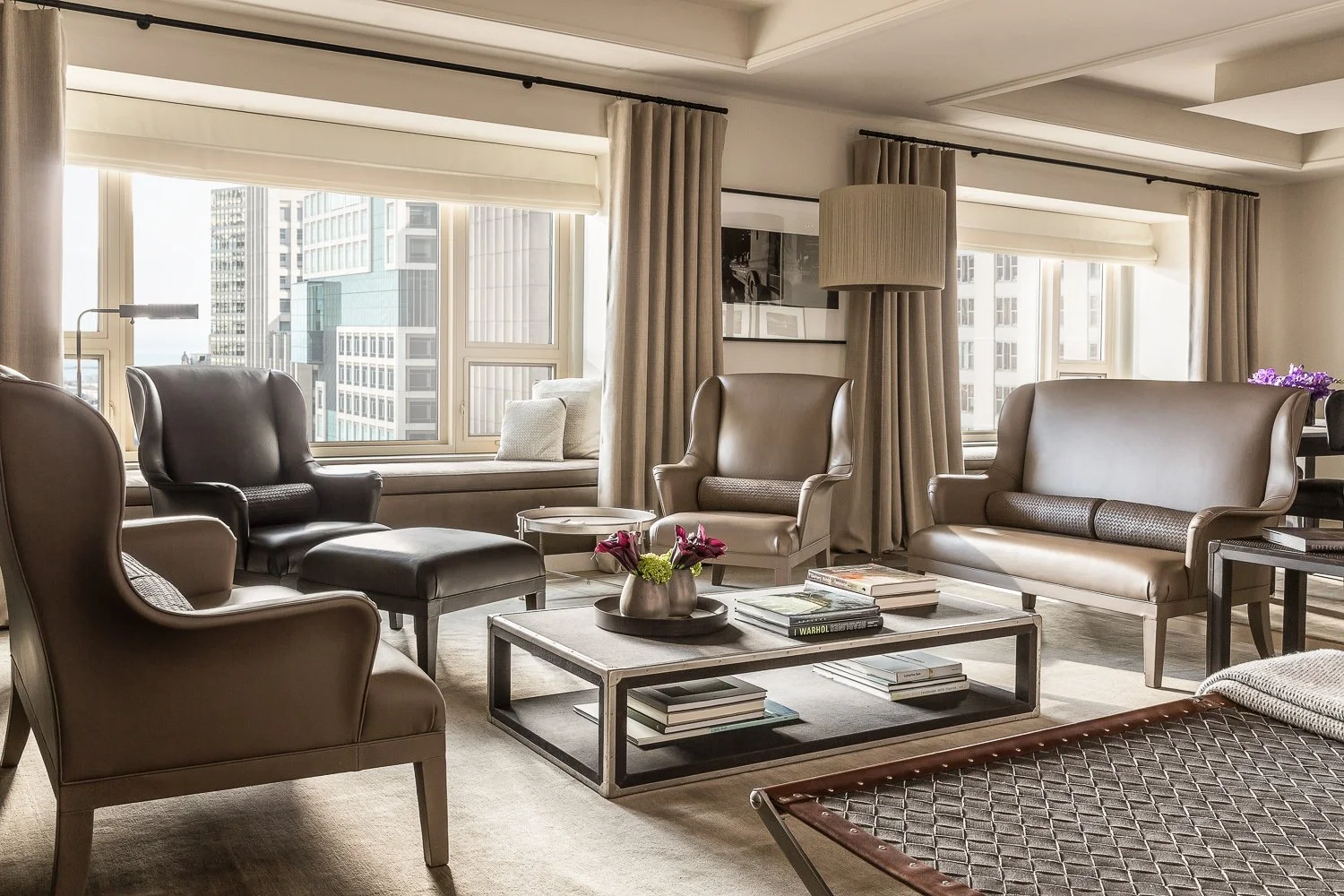 Modern living room with beige and black leather chairs and sofas, a wooden coffee table with books and a flower vase, large windows with city skyline views, beige curtains, and a neutral color palette.