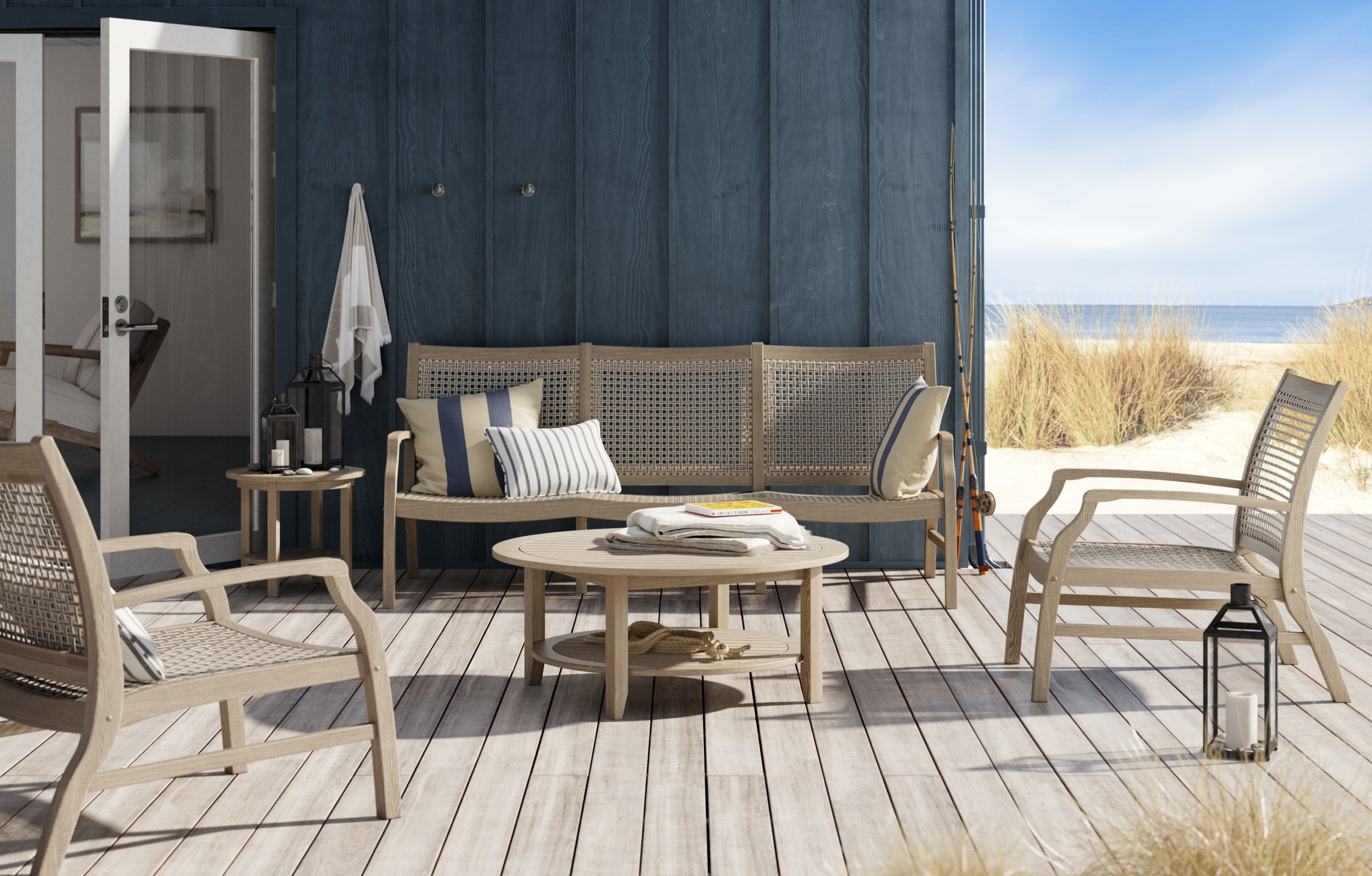 Beachside patio with wooden furniture, blue wall backdrop, overlooking sandy beach and ocean, decorated with pillows, lanterns, and fishing rods.