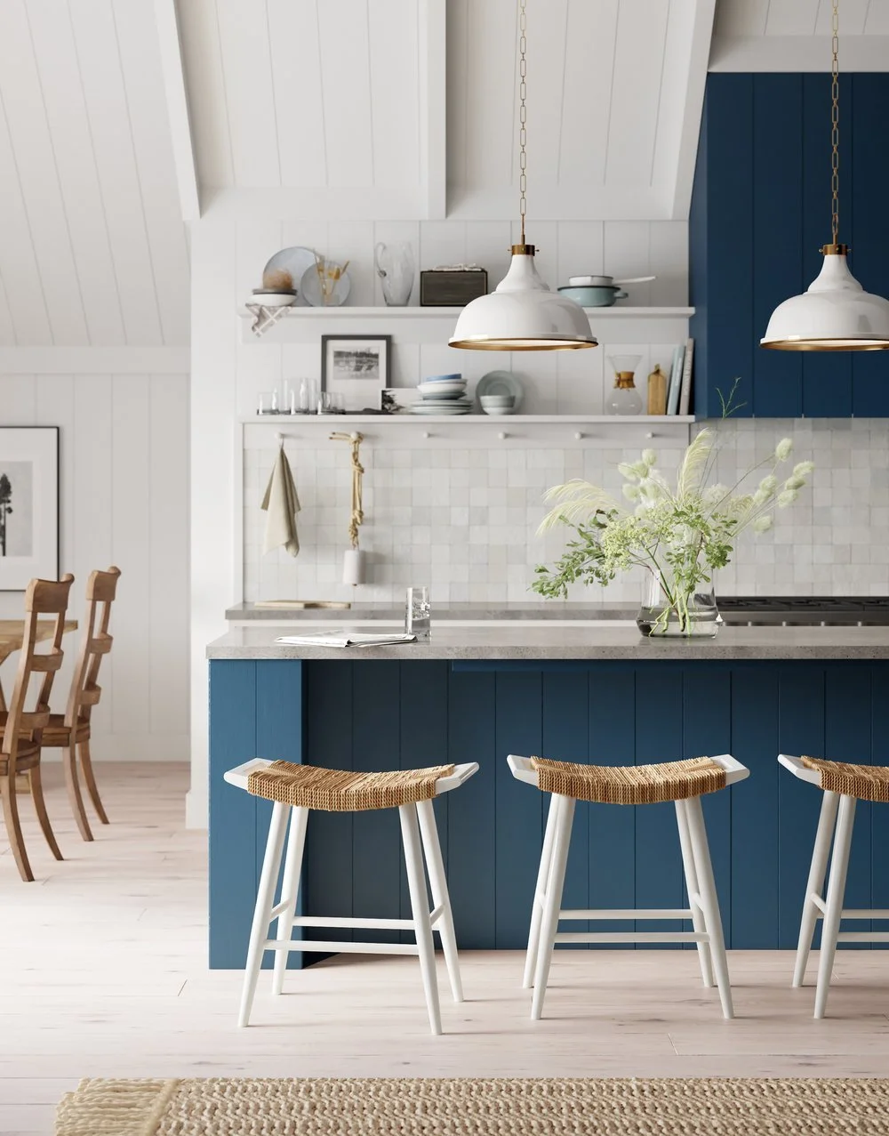 Modern kitchen with blue cabinetry, white walls, and a marble countertop. There are three white stools with woven seats at the counter, hanging white pendant lights, a vase with flowers, and open shelves with dishes and decor.
