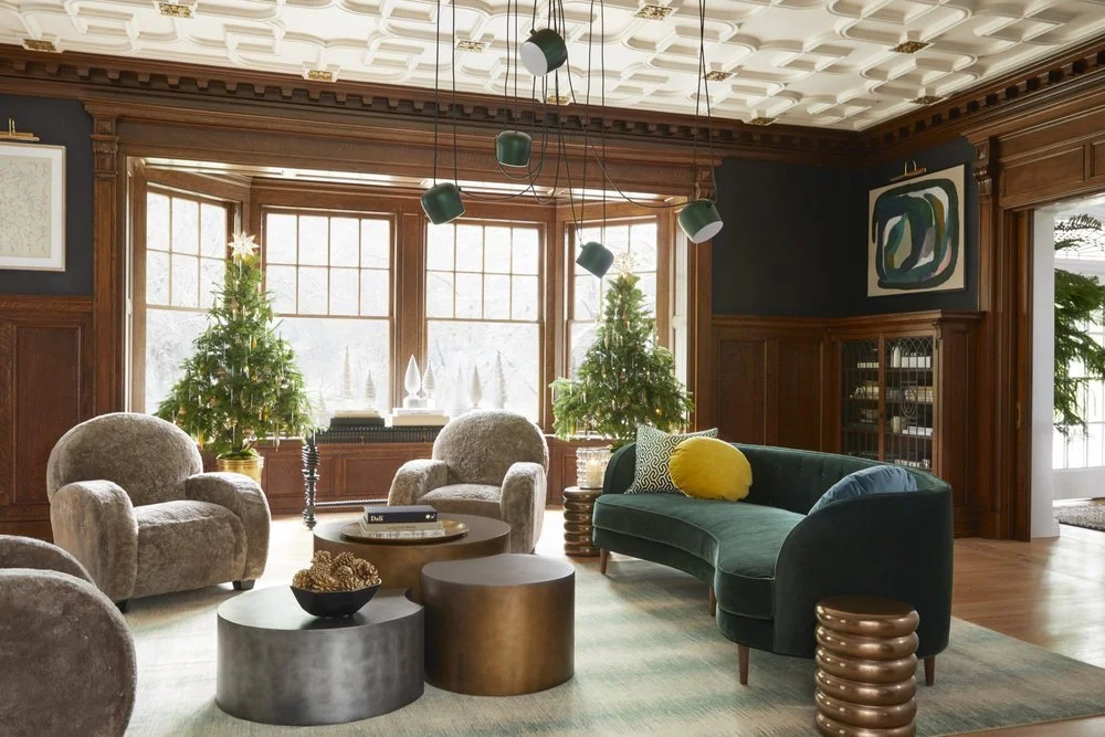 Living room with dark wood paneling, three Christmas trees, large bay windows, green velvet sofa, two plush armchairs, modern abstract wall art, and hanging pendant lights.