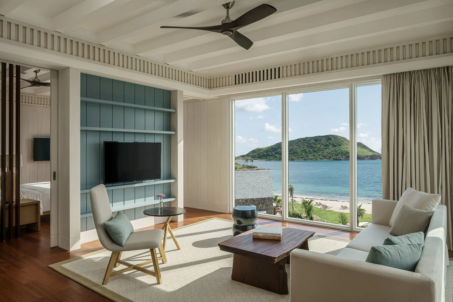 Living room with large glass windows overlooking a beach and green hill, white sofa, wooden coffee table, armchair, TV on a blue accent wall, and curtains pulled to the side.
