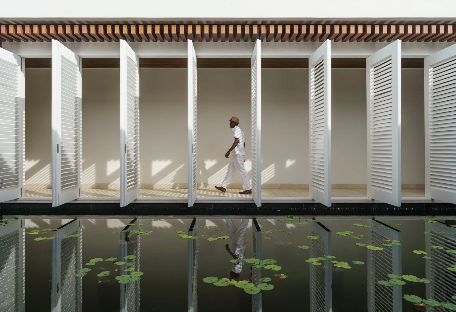 A man dressed in white walking past white open louvered doors with a pond with lily pads in the foreground, creating reflections of the doors and man.