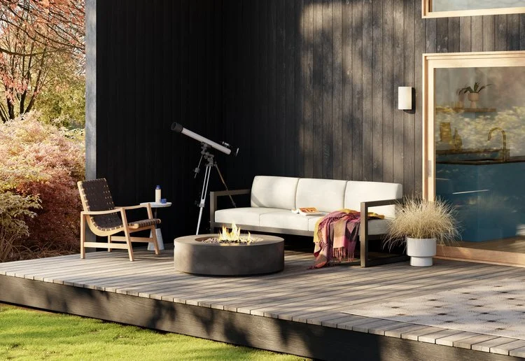 Outdoor patio with a white sofa, wooden armchair, telescope, fire pit, and potted plants next to a modern house with large windows.