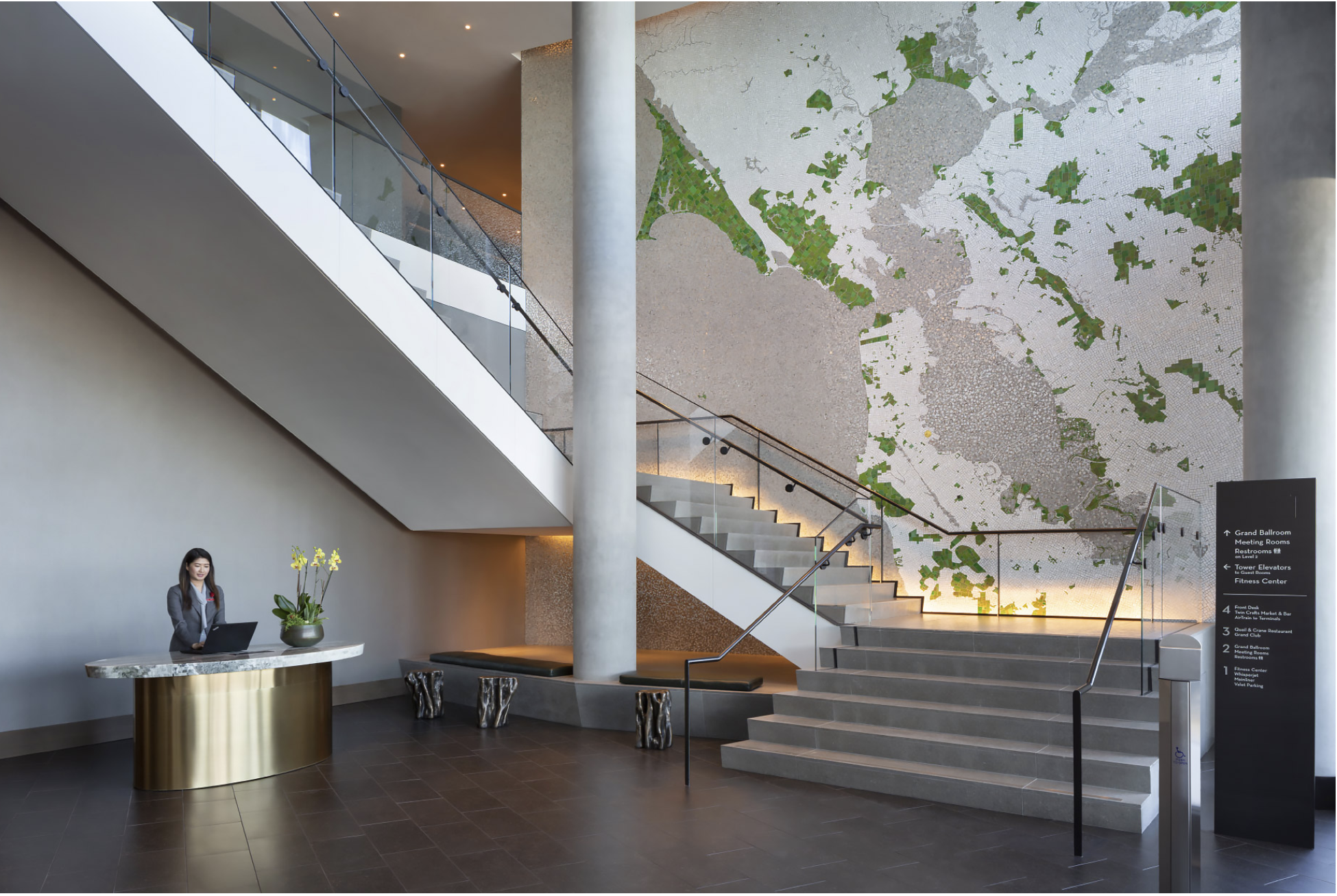 Interior of a modern hotel lobby with a woman at a reception desk, a large abstract world map on the wall, staircase with glass railing, and a black sign with white text providing directions.