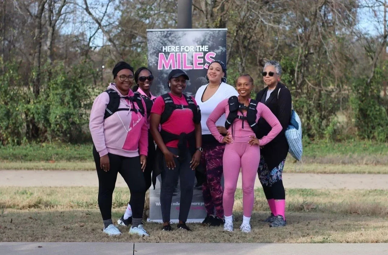 Group of six women standing outdoors, dressed in activewear, smiling, with a banner behind them reading 'Here for the Miles', trees in the background.