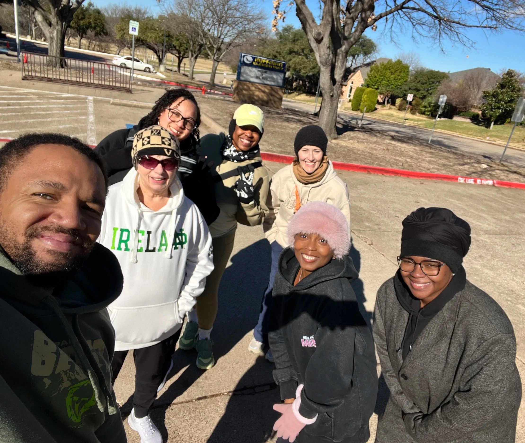 Group of eight diverse people smiling outdoors on a sunny day, standing on a parking lot with bare trees and a few buildings in the background.