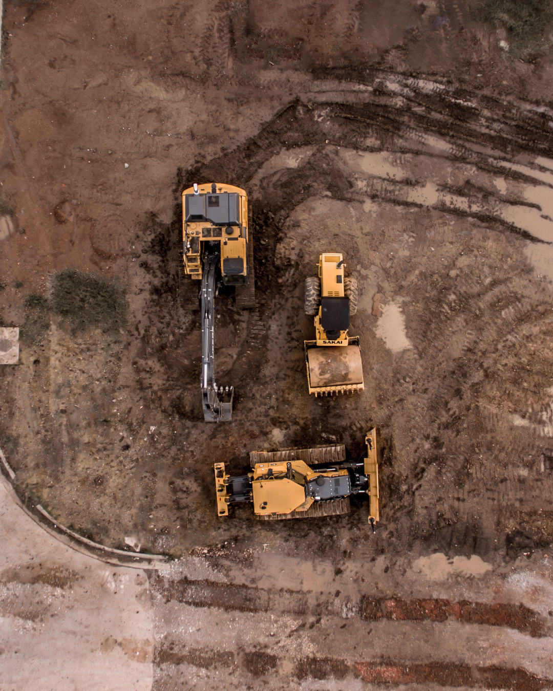 Aerial view of a civil construction site. There are three machines visible working on the ground. An excavator, roller and dozer..
