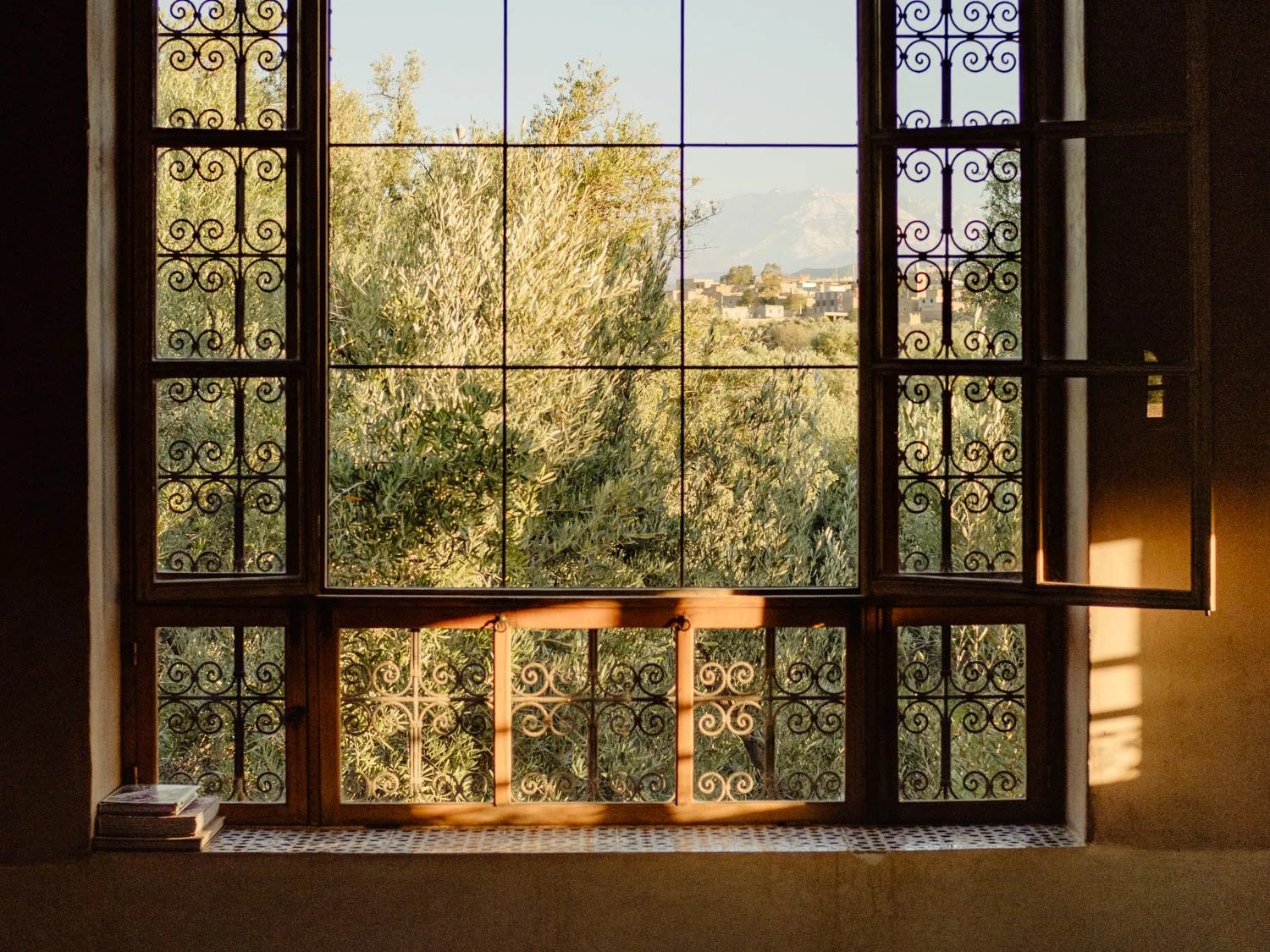 Interior view through a Moroccan window framing olive trees and a garden at sunset, with warm light crossing tiled surfaces and architectural edges.