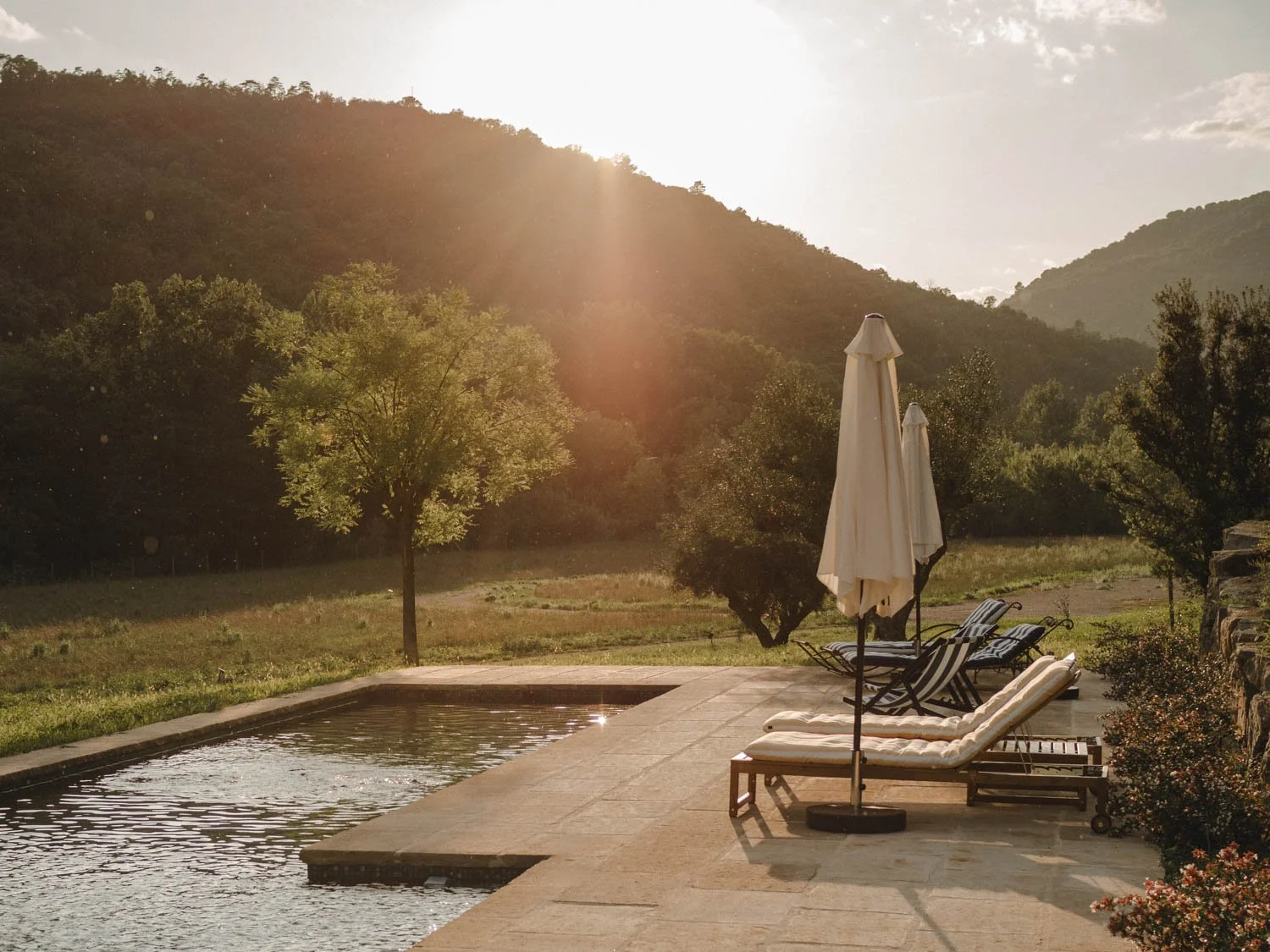 Natural pool at sunset with two empty hammocks and closed umbrellas as light rain falls over the landscape under fading light.
