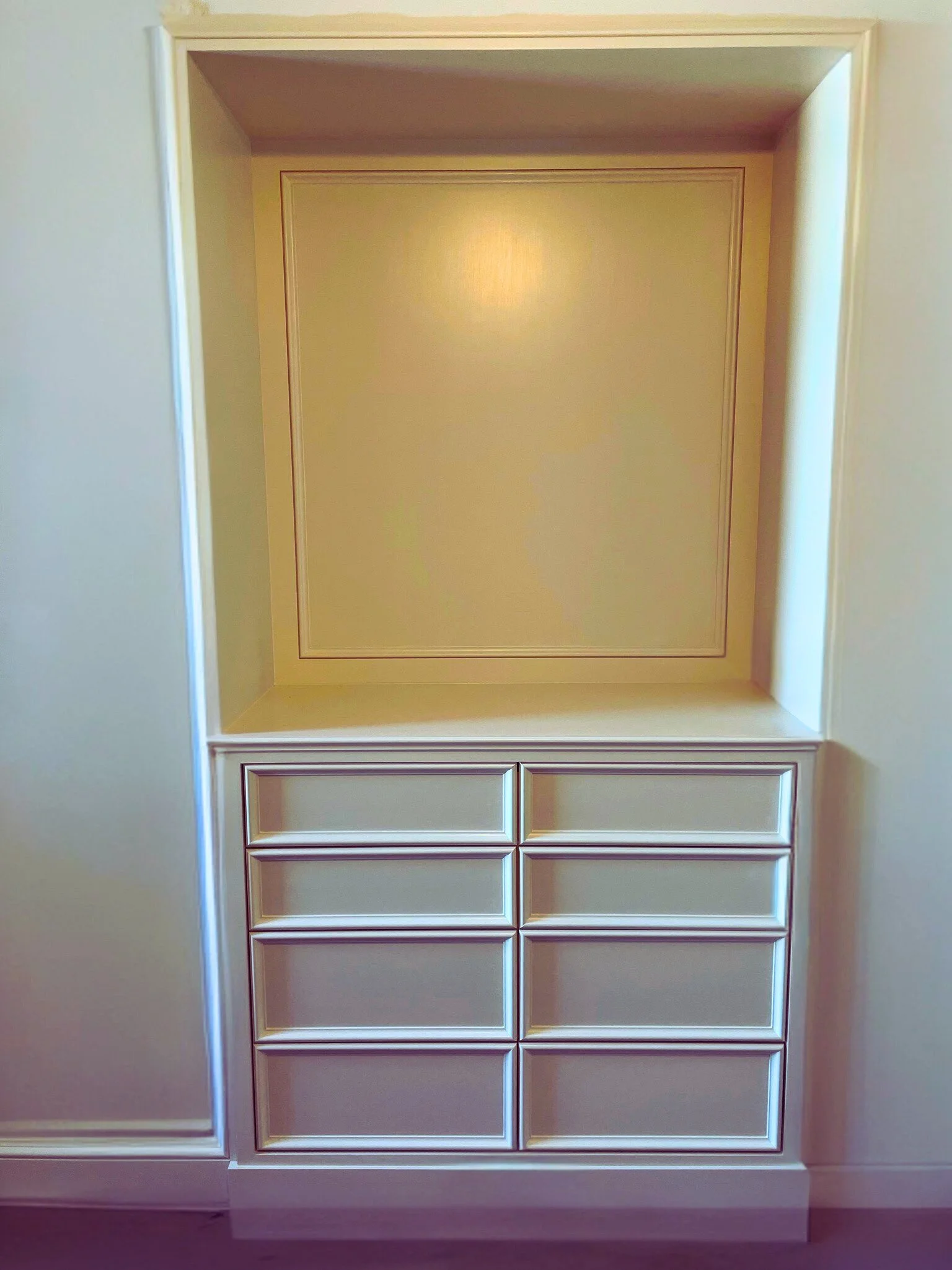 Built-in cream-colored cabinet with nine open drawers below a large open square space, set in a wall with decorative molding.