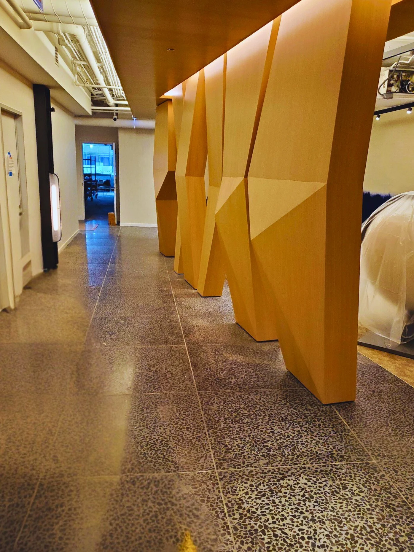 Interior hallway with wooden geometric partition, terrazzo floor, and visible ceiling pipes.