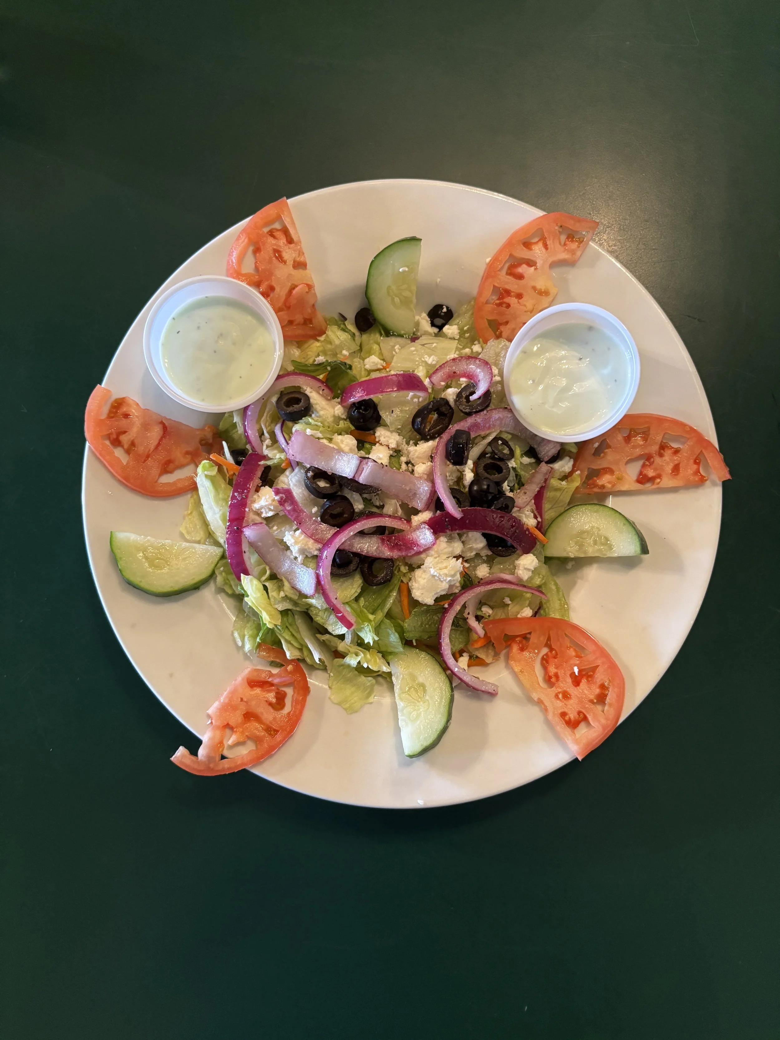 Fresh garden salad with lettuce, tomato slices, cucumber slices, red onion, black olives, feta cheese, and two small containers of dressing on a white plate on a dark green surface.
