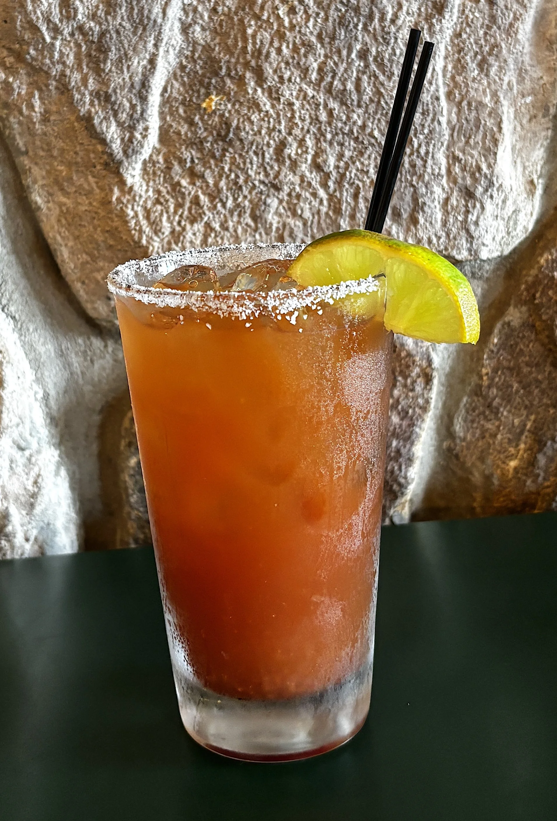 A tall glass of iced tea garnished with a lemon wedge, with a salted rim and two black straws, against a stone brick background.