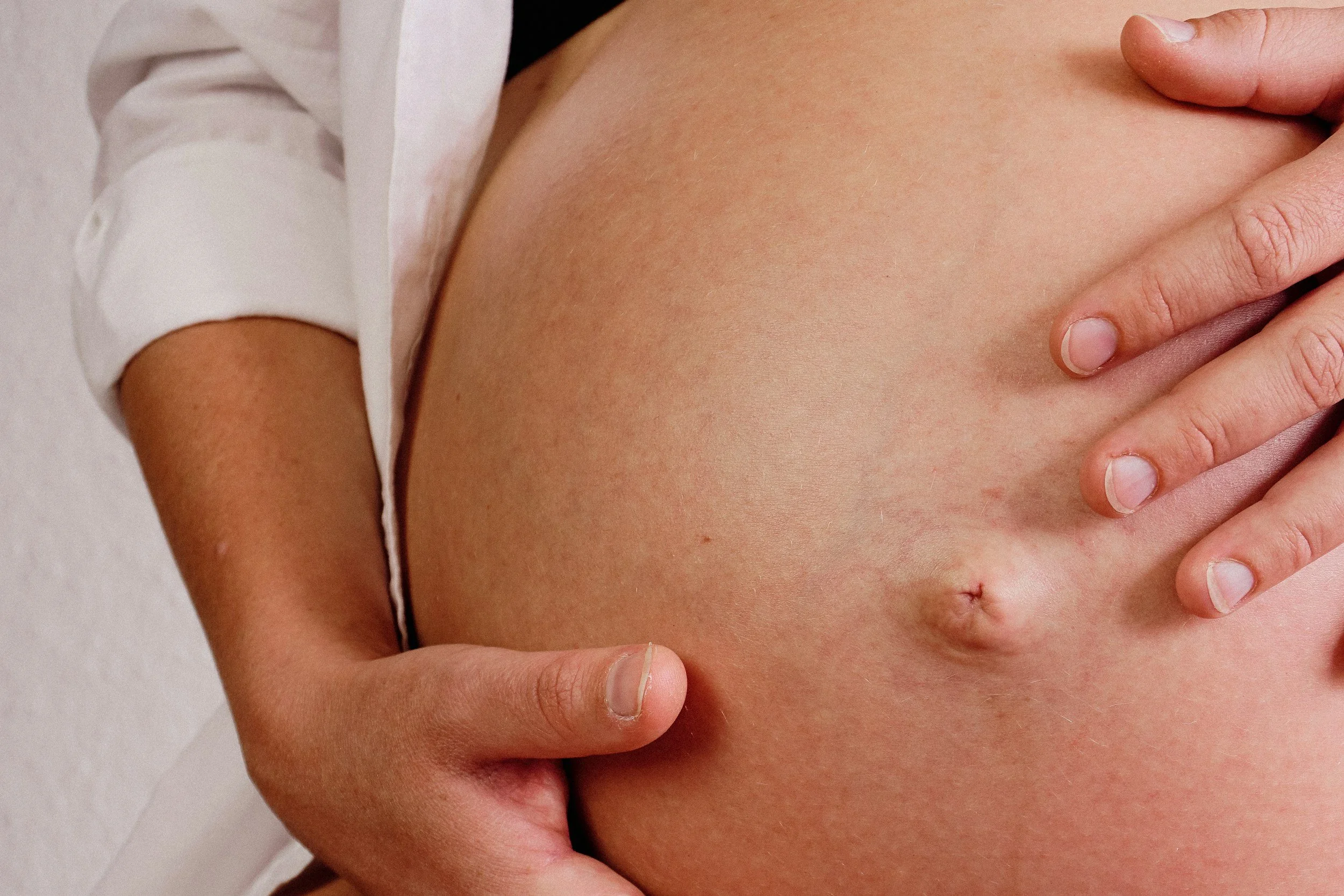 Close-up of a pregnant woman's belly, with one hand on her stomach and the other hand resting on her side, wearing a white shirt with the sleeve rolled up.