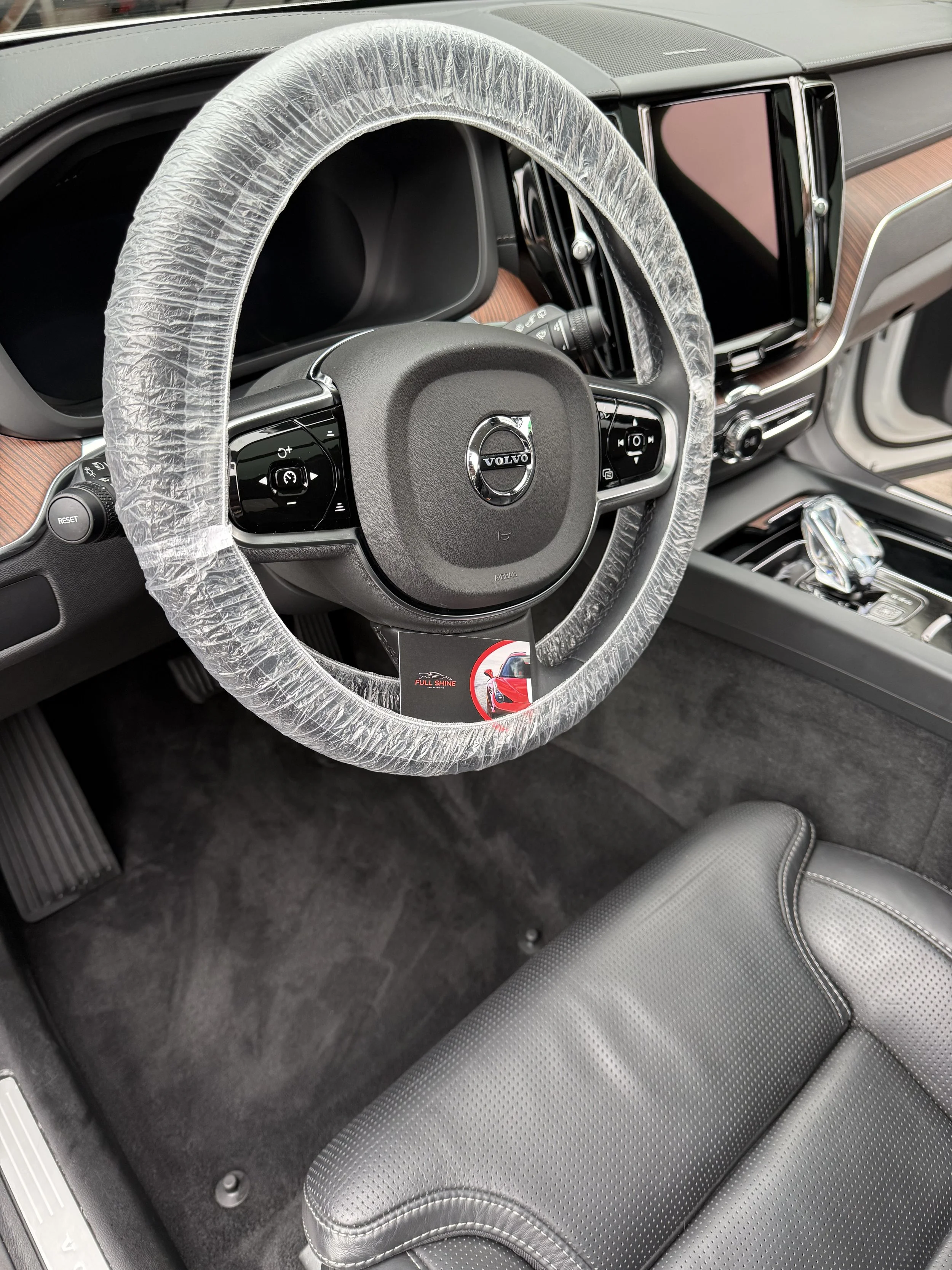 Interior of a Volvo vehicle, showing a steering wheel with a protective cover, digital dashboard, infotainment screen, gear shift, and black leather seats.