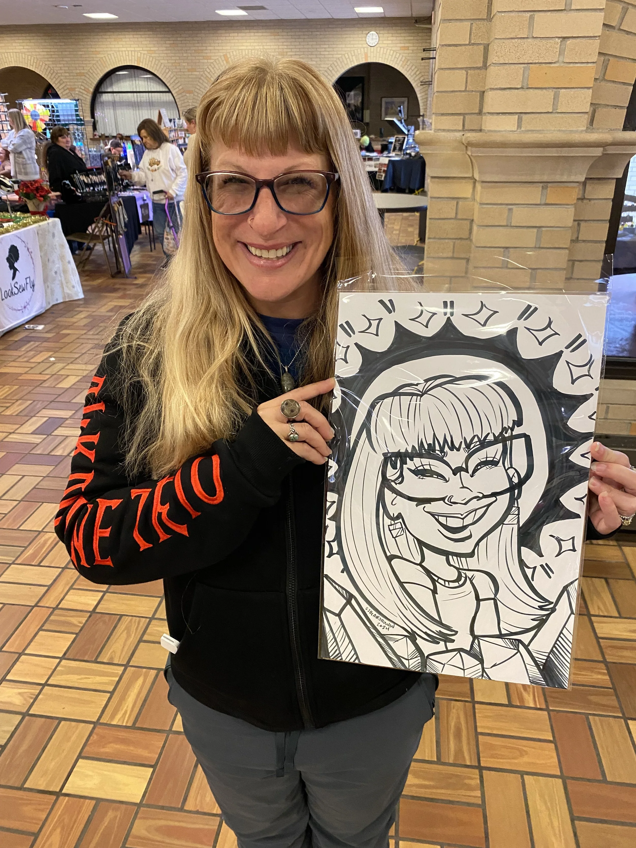 A woman with long blonde hair, glasses, and a black jacket smiling and holding a framed black-and-white caricature of herself in an indoor market or craft fair setting.