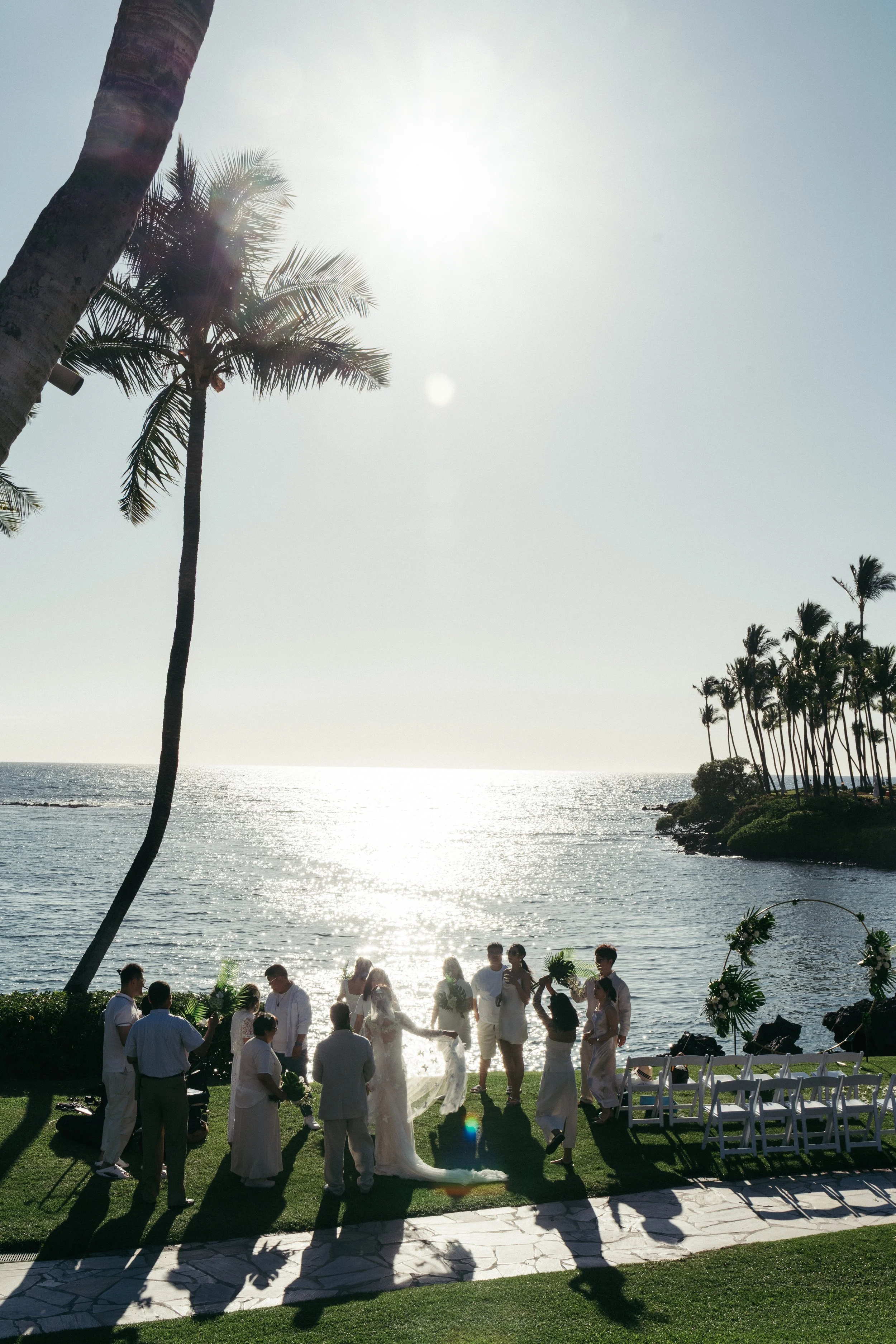 A wedding ceremony taking place outdoors on a grassy area by the ocean, with palm trees and the sun in the sky, and guests and the bride and groom in white attire.