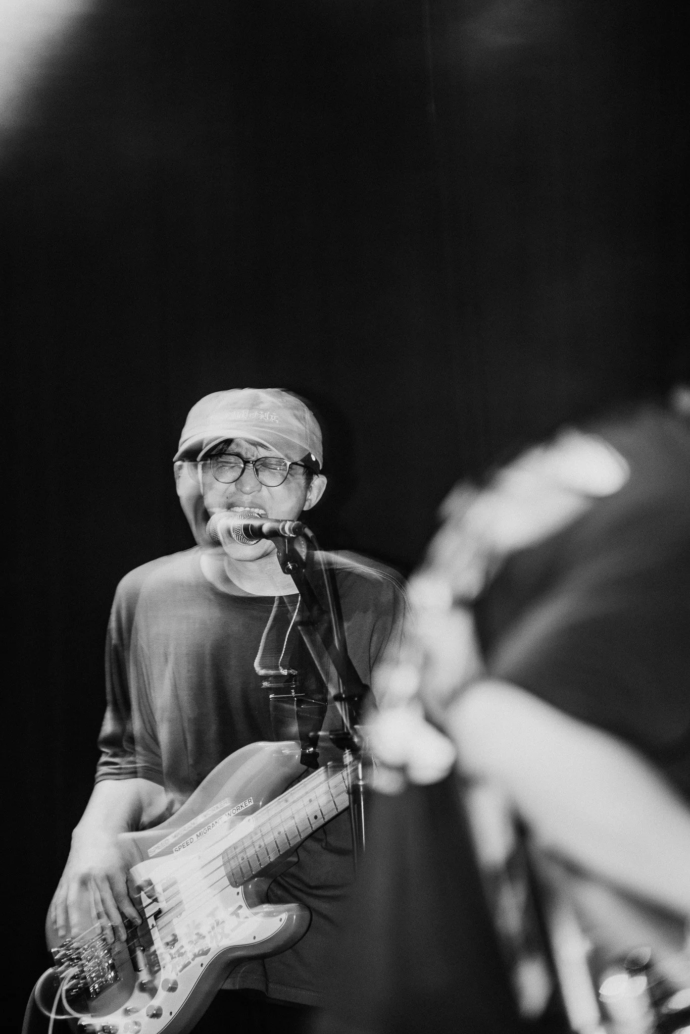 A person singing into a microphone and playing an electric bass guitar on stage, with a dark background.