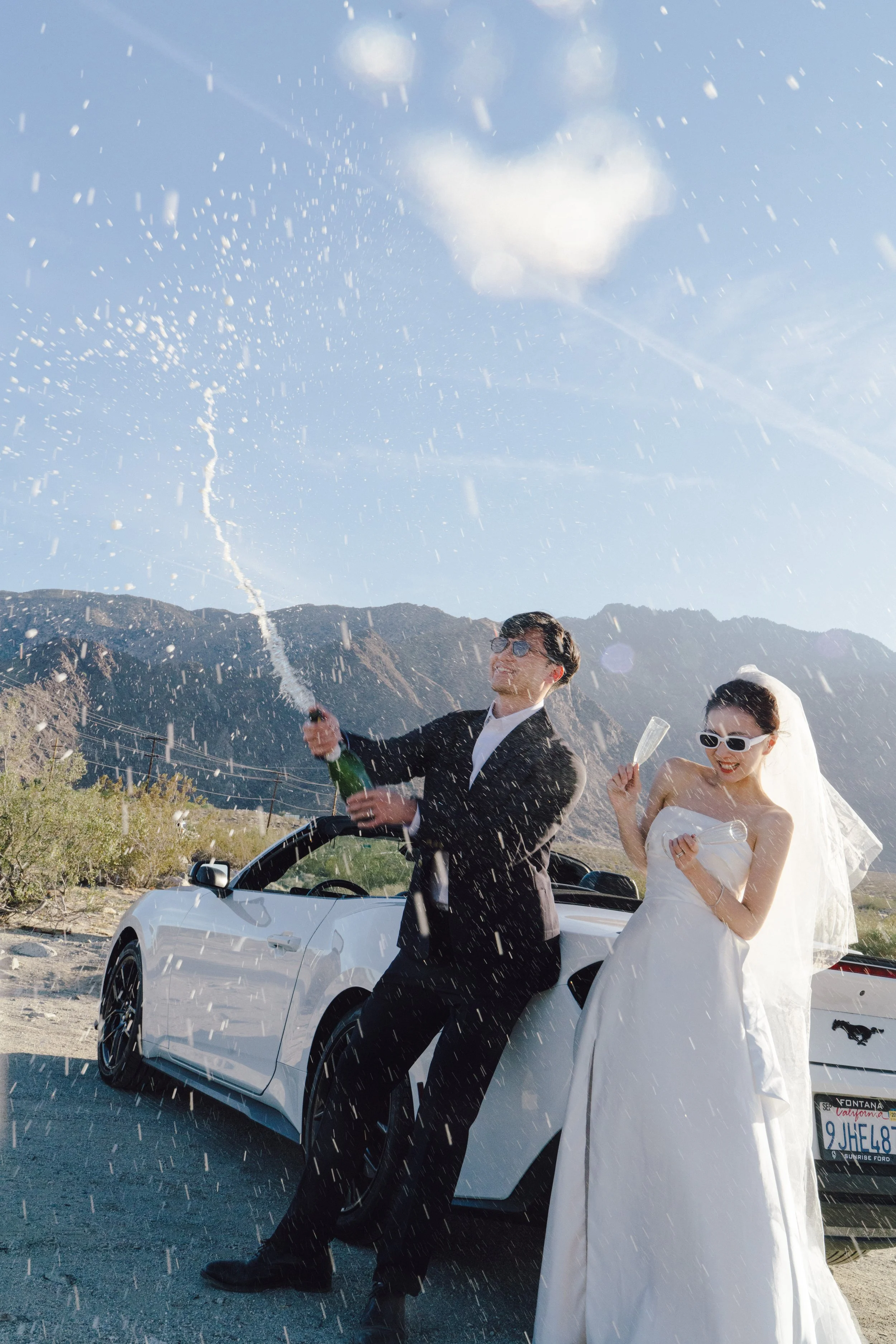 A newlywed couple in wedding attire celebrating outdoors, opening champagne near a white convertible car with mountains in the background on a sunny day.
