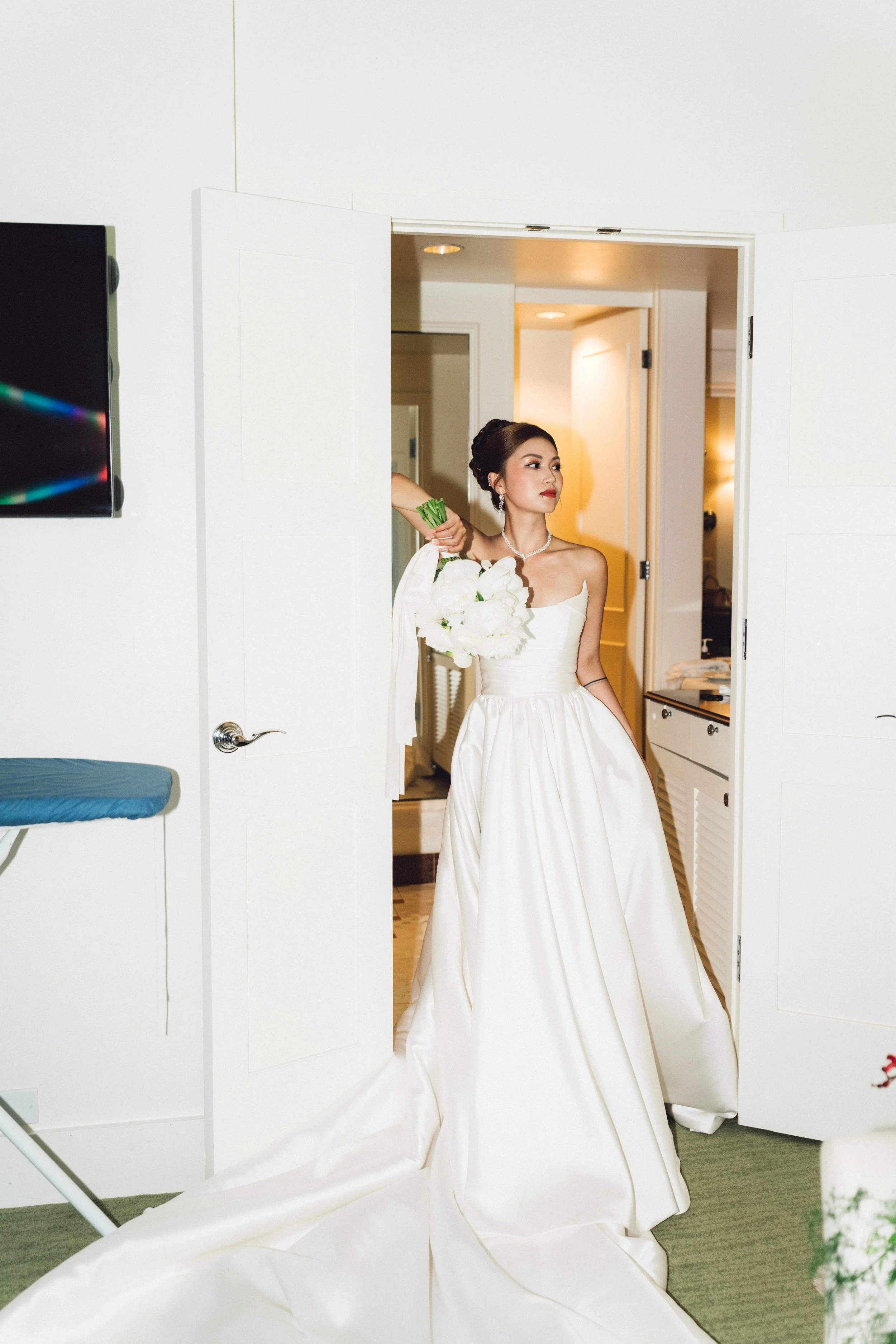 A bride in a white wedding gown holding a bouquet of white flowers, standing in a hotel room doorway, with a pensive expression.