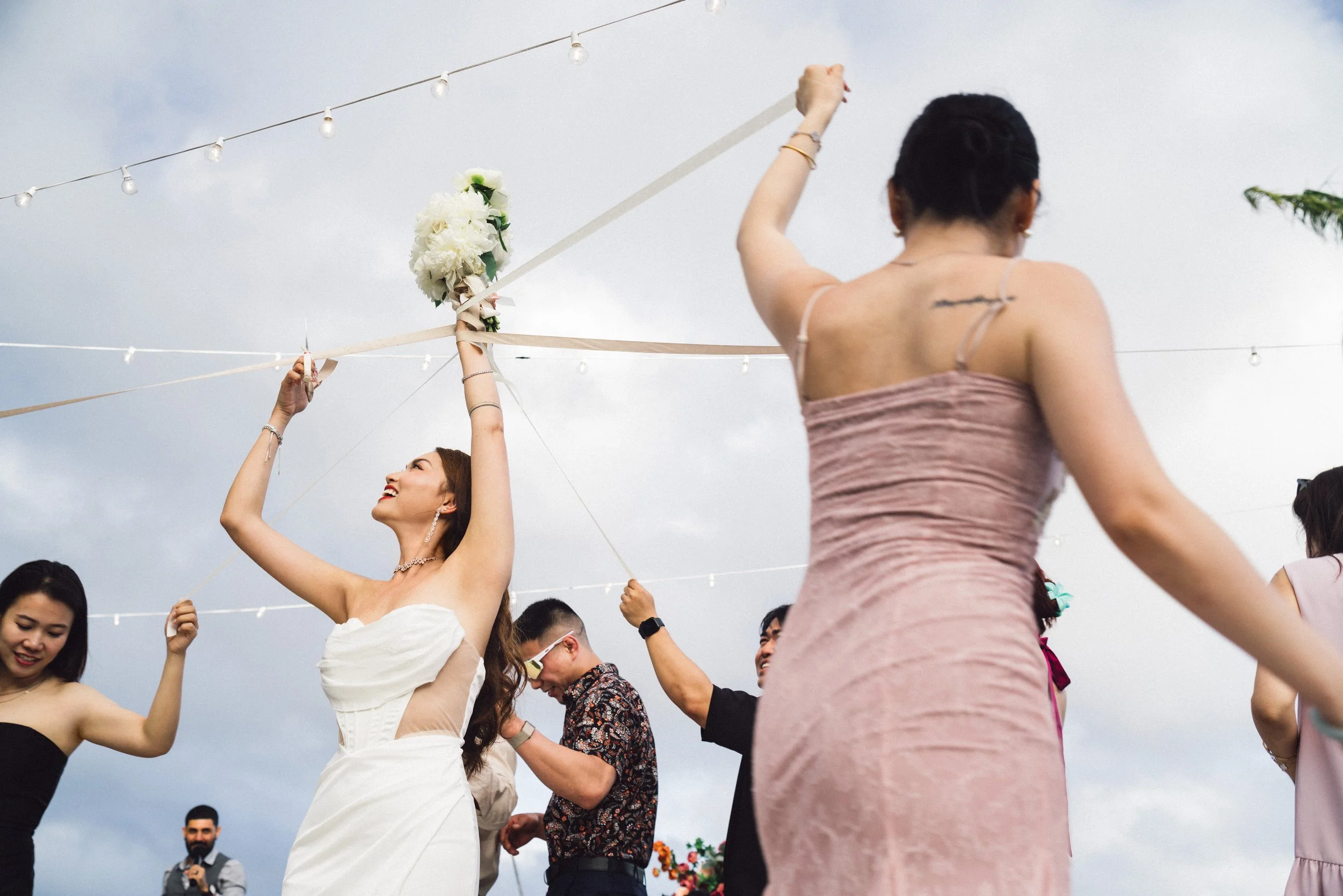 People celebrating at an outdoor wedding reception, with a woman in a white dress holding a bouquet and others dancing under string lights.