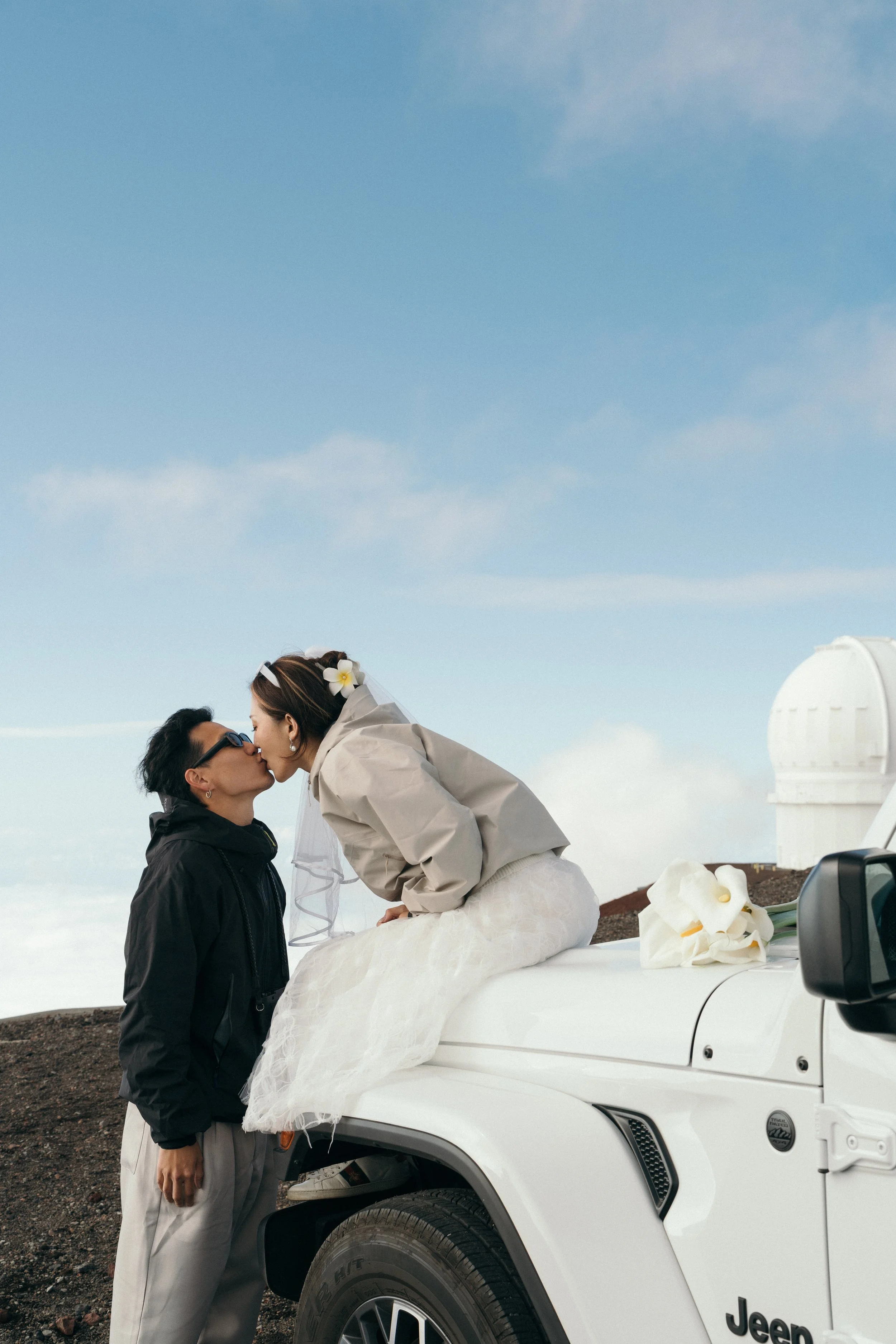 A couple sharing a kiss during their wedding, with the bride sitting on the hood of a white Jeep vehicle on a rocky terrain under a blue sky.