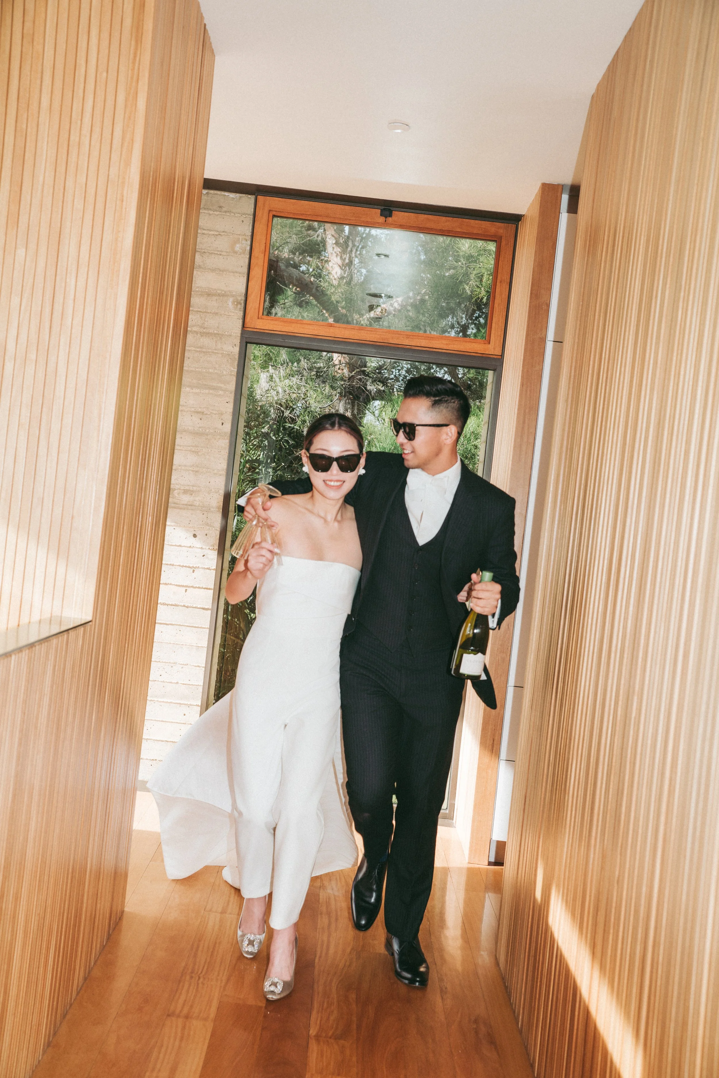 A happy couple, dressed in wedding attire, walks arm-in-arm through a wooden hallway, with the man holding a bottle of champagne. Both wear sunglasses and are smiling.
