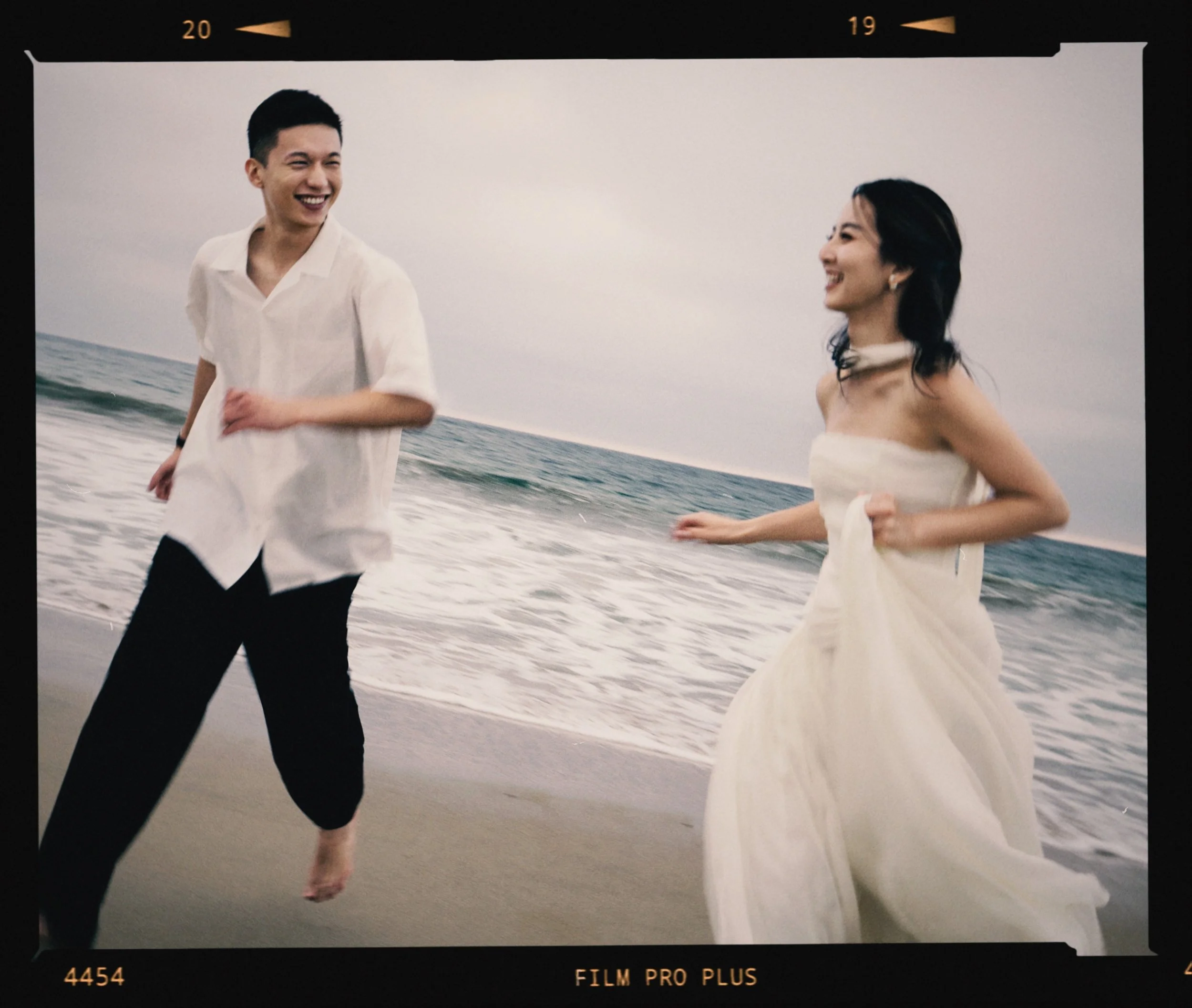 A young man and woman are running along the beach near the ocean, smiling and enjoying the moment; the woman is wearing a strapless white dress, and the man is dressed casually in a white shirt and black pants.