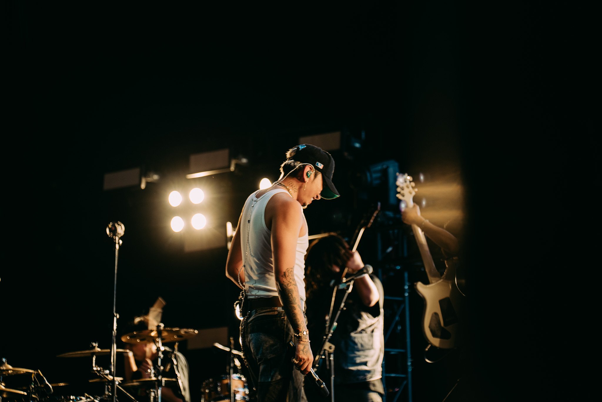 A musical band performing on stage with a lead singer wearing a black cap and sleeveless shirt, holding a microphone, under bright stage lights.