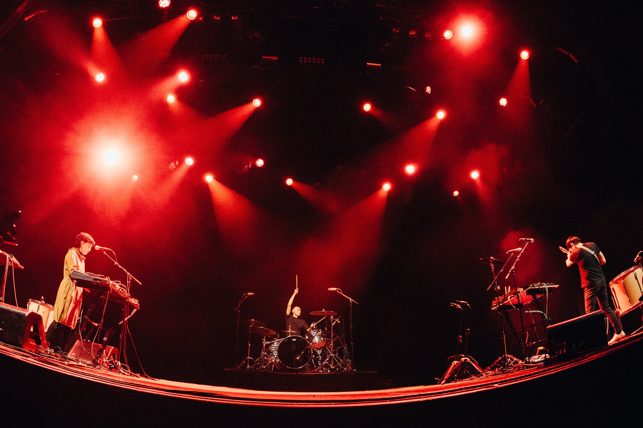 A band performing on stage with red lighting, featuring a keyboardist, a drummer, and a guitarist, with microphones and musical equipment.