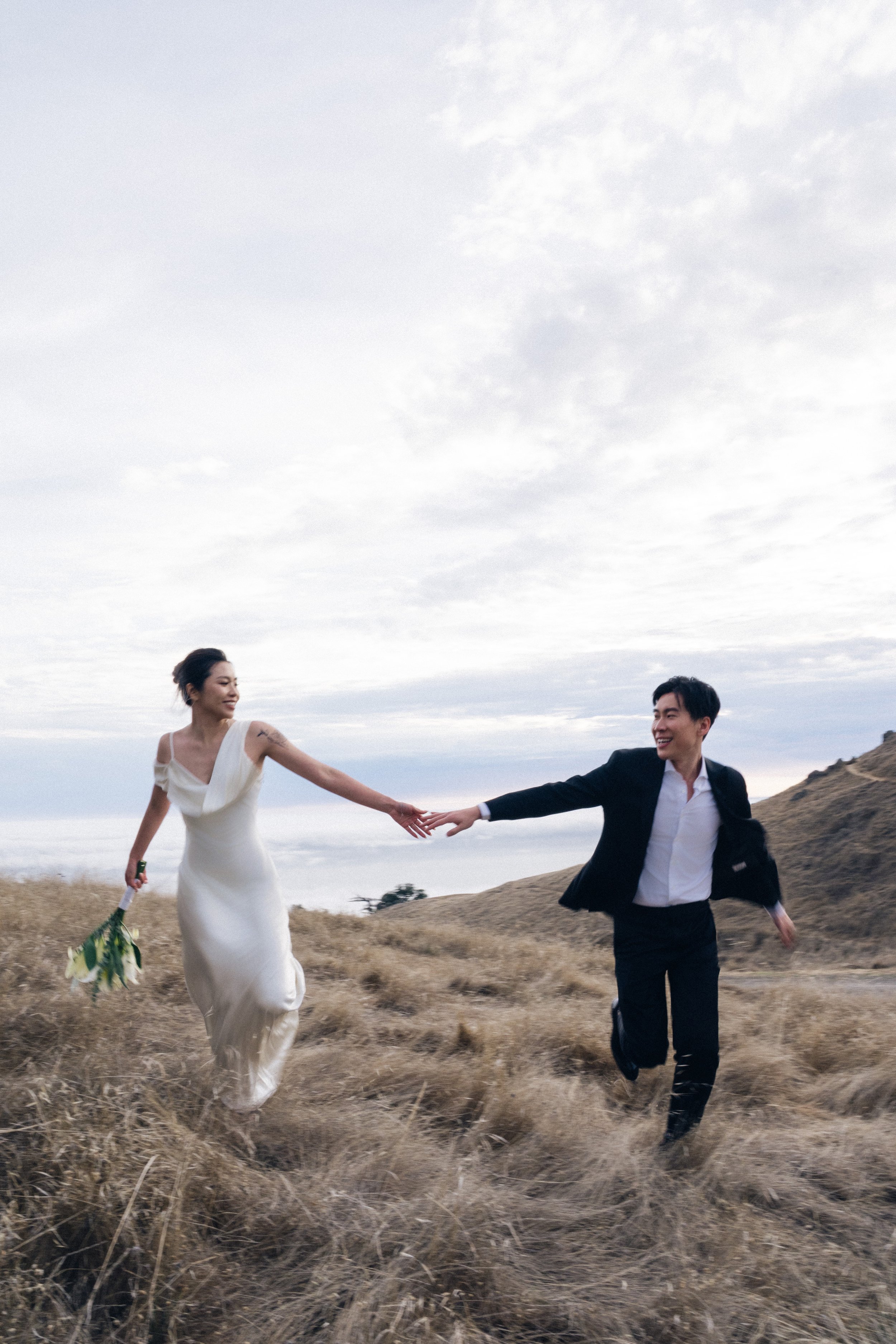 A woman in a white dress holding a bouquet running hand-in-hand with a man in a black suit on a grassy hill under a cloudy sky.