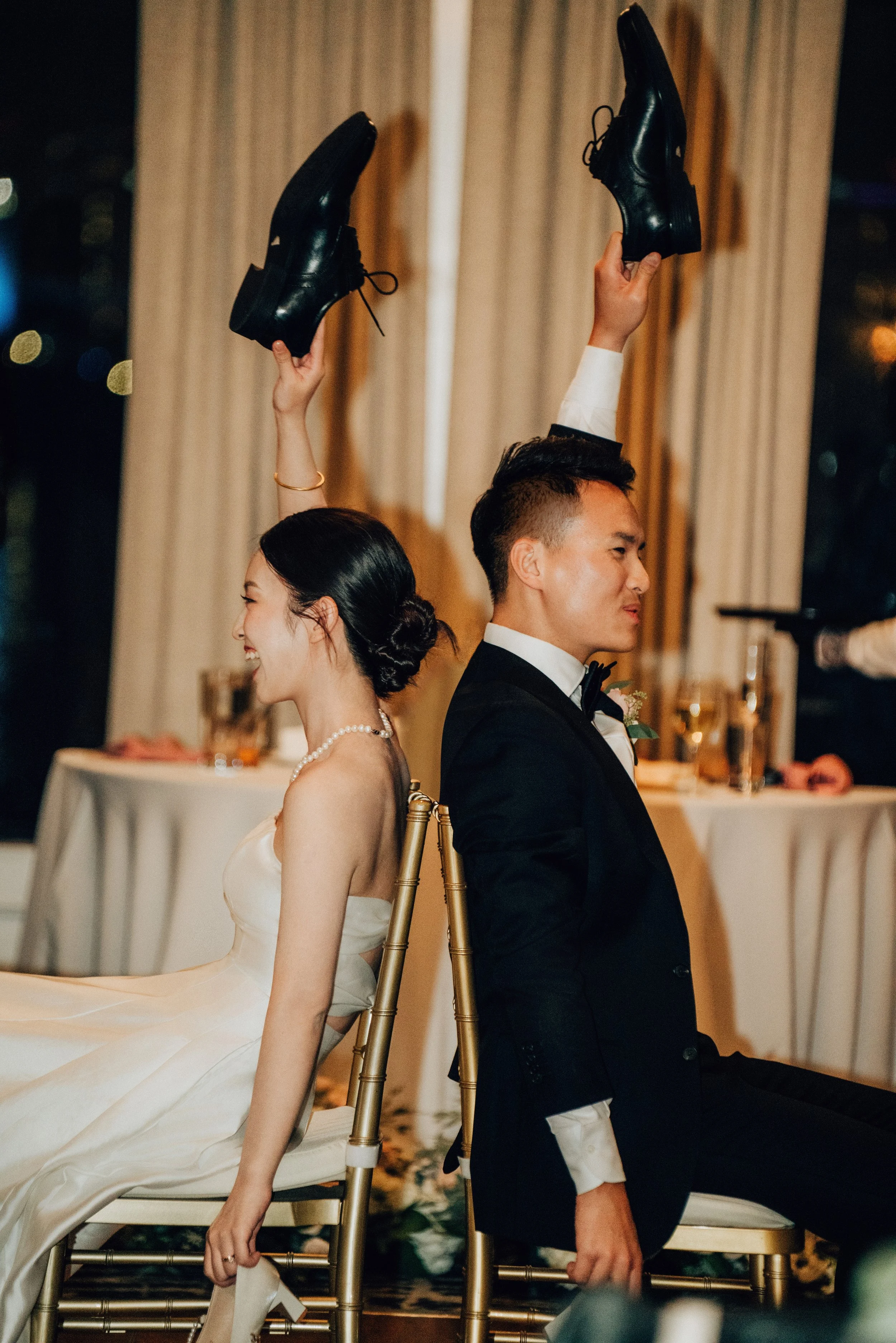 Bride and groom sitting back-to-back on chairs during their wedding reception, playing a game with shoes raised in the air.