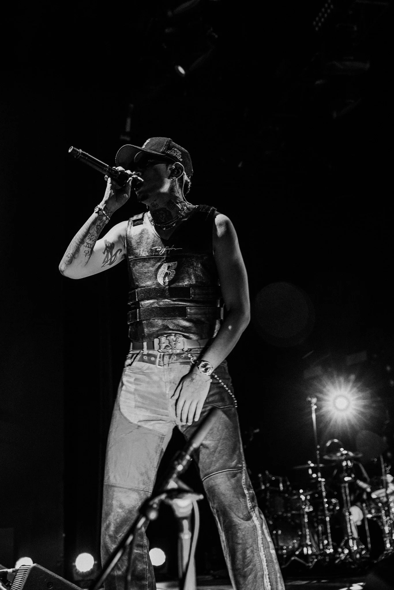 A black and white photo of a performer on stage, singing into a microphone, wearing a baseball cap, sleeveless vest, baggy jeans, and jewelry, with stage lights behind and a drum set visible in the background.