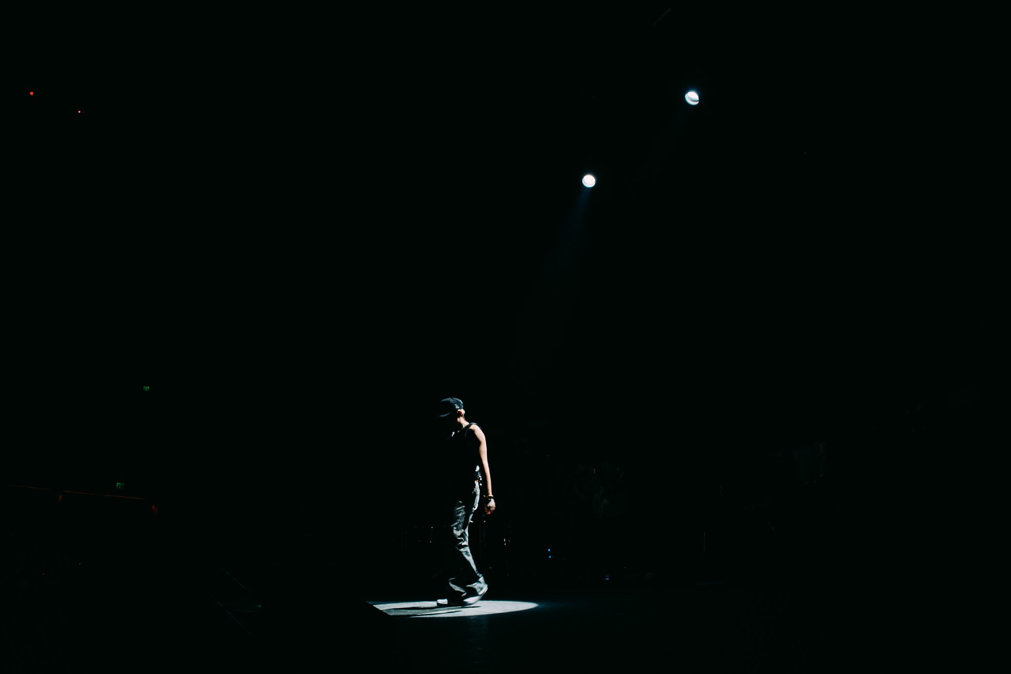 A performer standing alone in a spotlight on a dark stage with two bright lights shining from above.