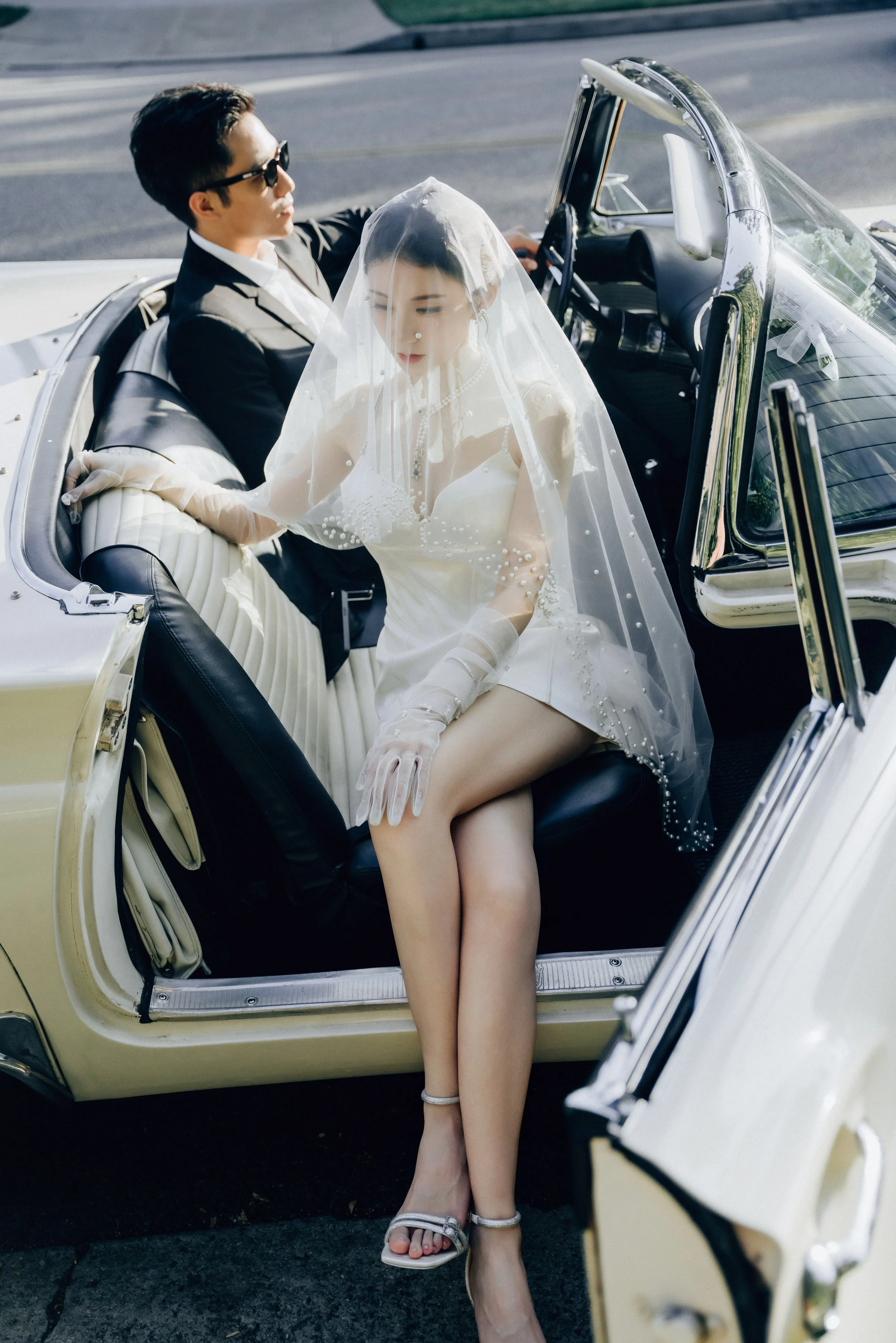 A bride in a white wedding dress and veil sitting in a vintage convertible car with a groom in a tuxedo, on a street.