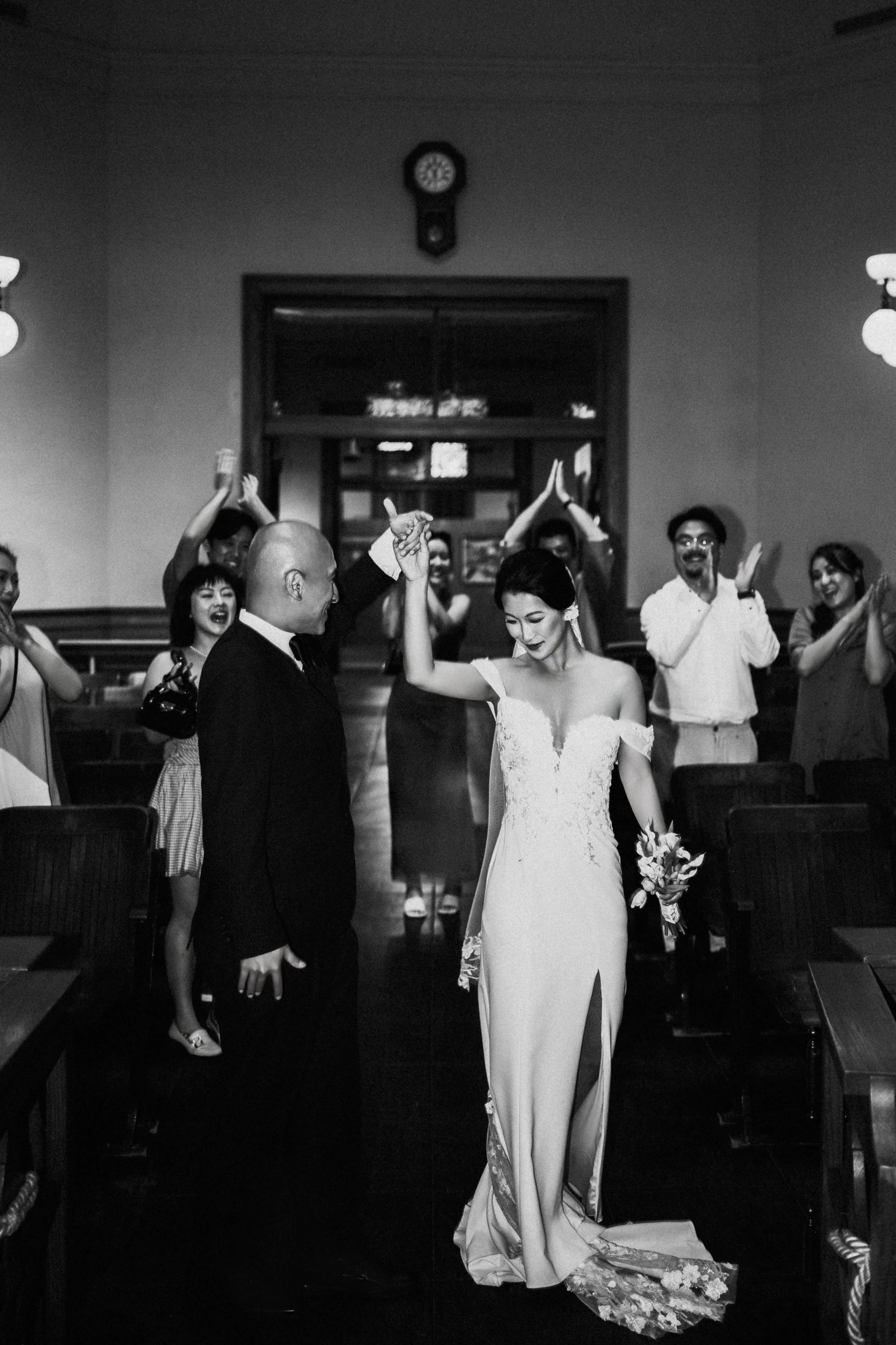 A black and white photo of a wedding scene with a bride and groom dancing, surrounded by their guests clapping and celebrating in a decorated indoor venue.