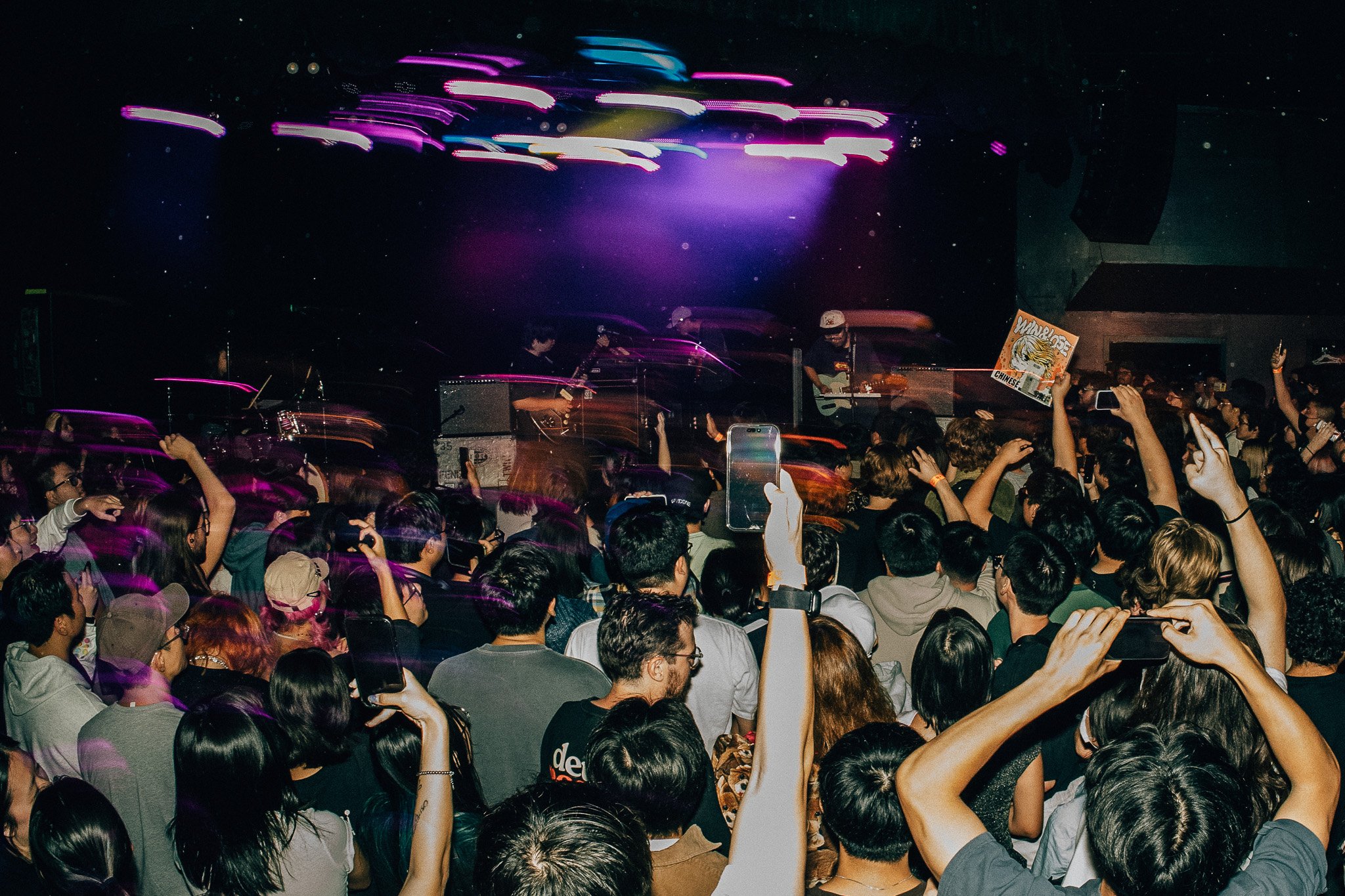 A large crowd of people watching a live music concert in a dark indoor venue with purple and green stage lights. The band is performing on stage with guitars and drums. Audience members are raising their hands and taking photos.