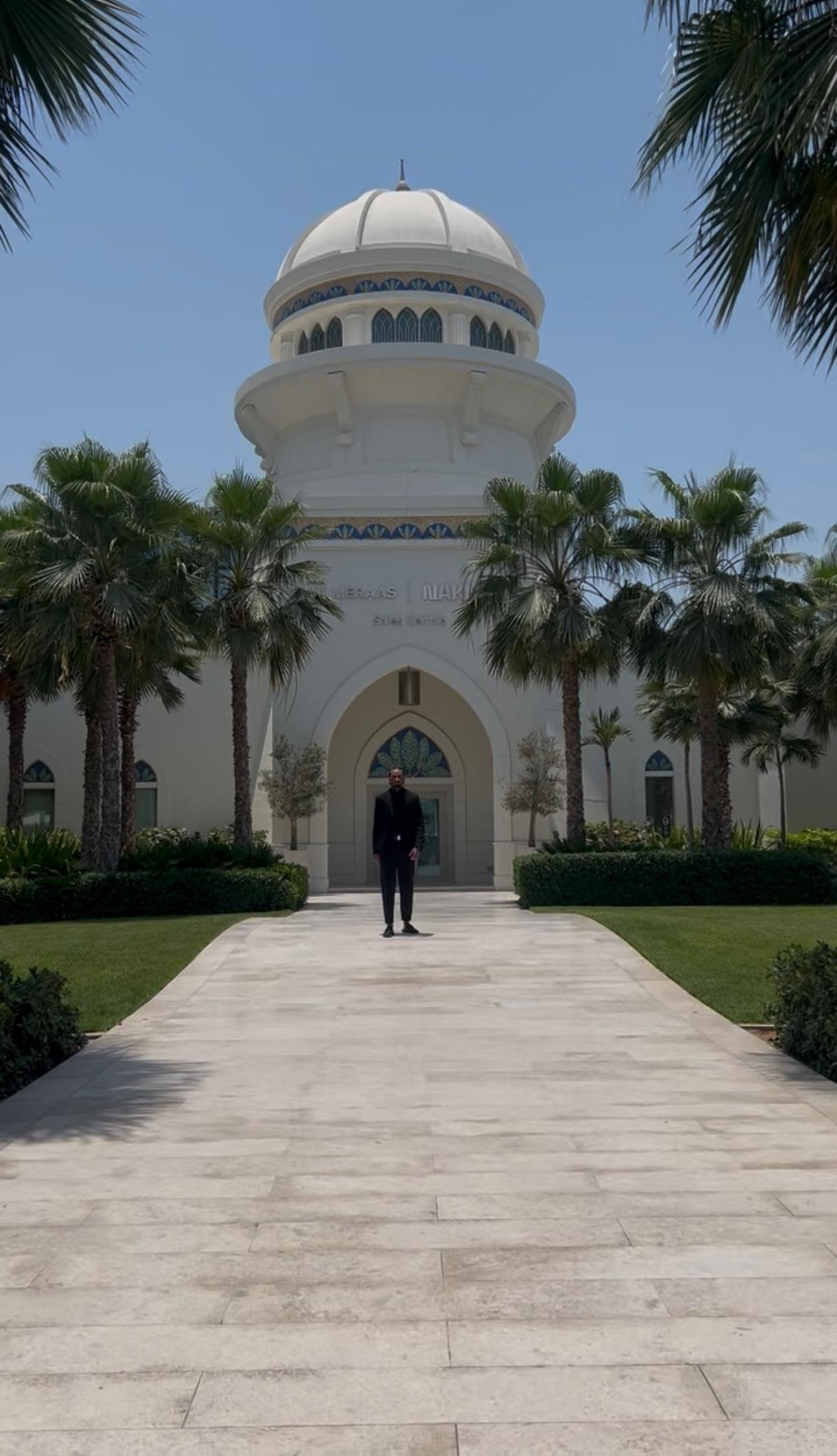 A man in a black suit standing on a wide, beige stone pathway leading to a white building with a dome, surrounded by palm trees under a clear blue sky.