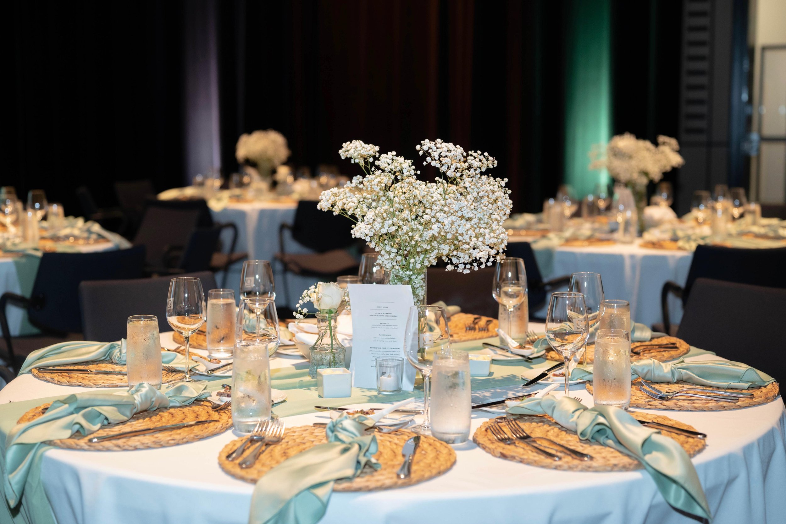 Elegant banquet table setting with a floral centerpiece, blue napkins, woven placemats, glasses, and cutlery at a decorated event.