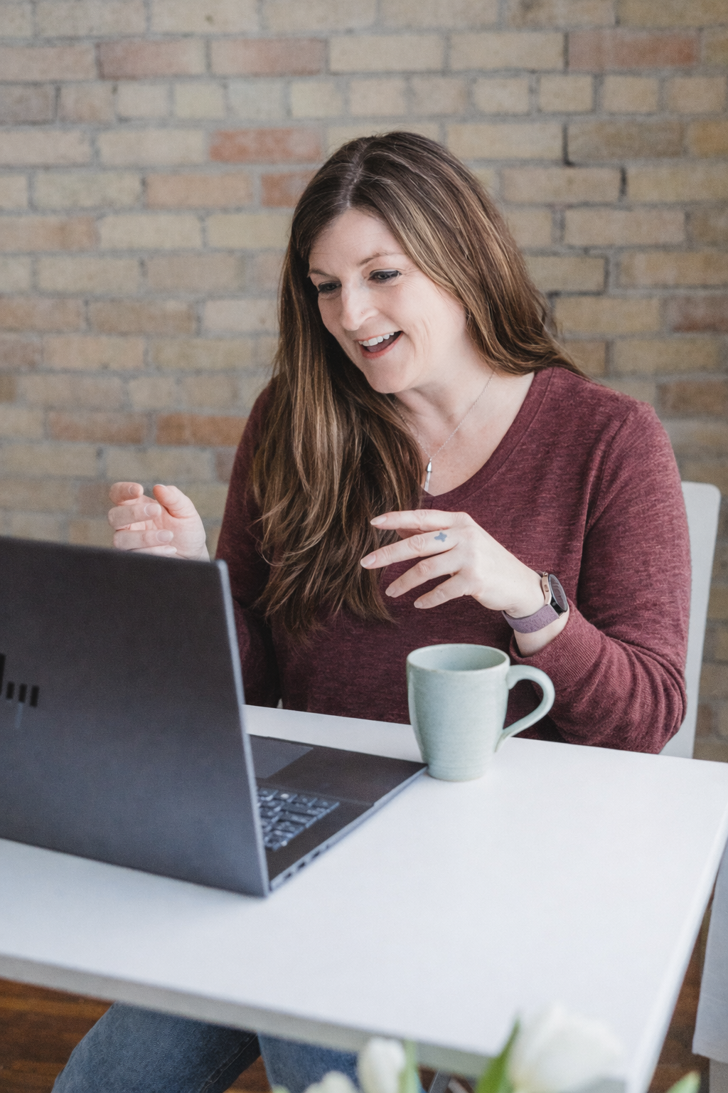 Telehealth mental health therapy with a woman with long brown hair wearing a maroon sweater and a smartwatch is sitting at a white table, looking at her laptop with a smile, beside a green mug, in front of a brick wall.